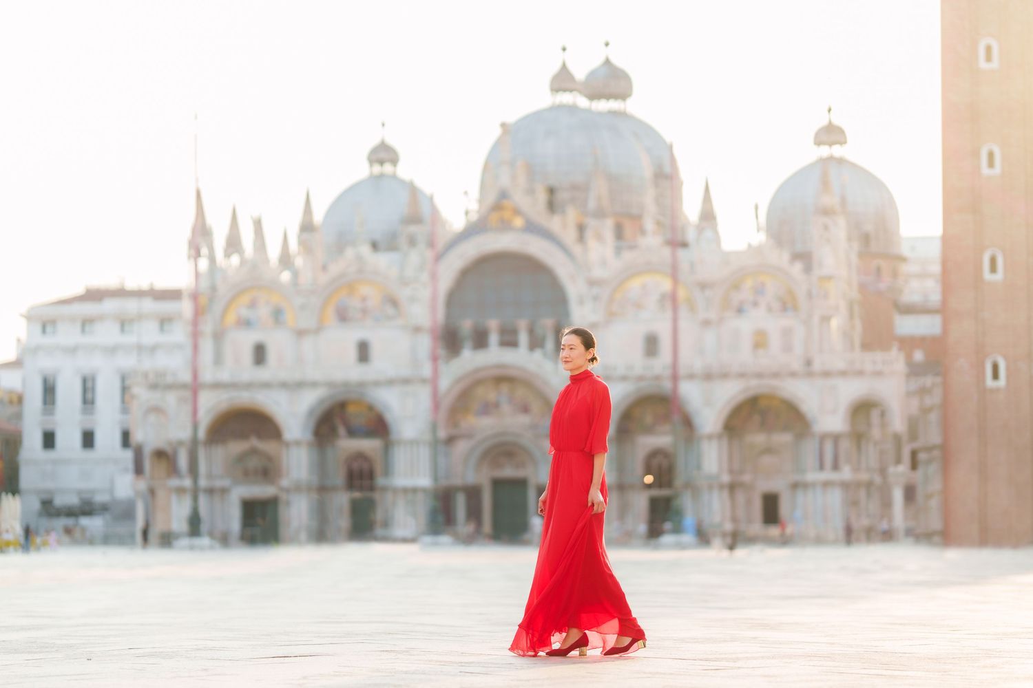 Woman in red gown walking in St. Mark's Square, Venice, with the iconic Basilica di San Marco in the background.