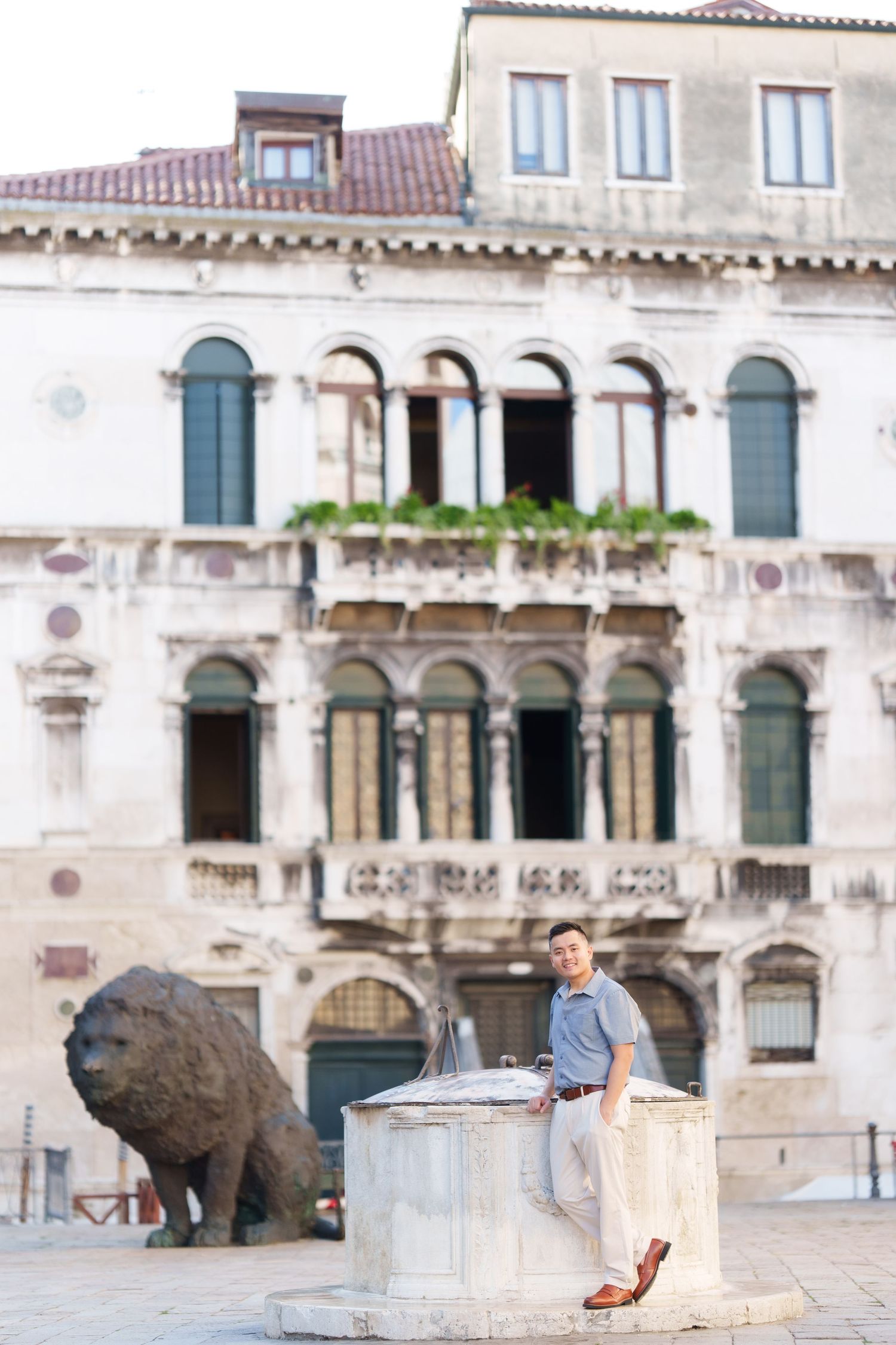 Man leaning on a stone well in a historic Italian piazza with a bronze lion sculpture and ornate Renaissance building.