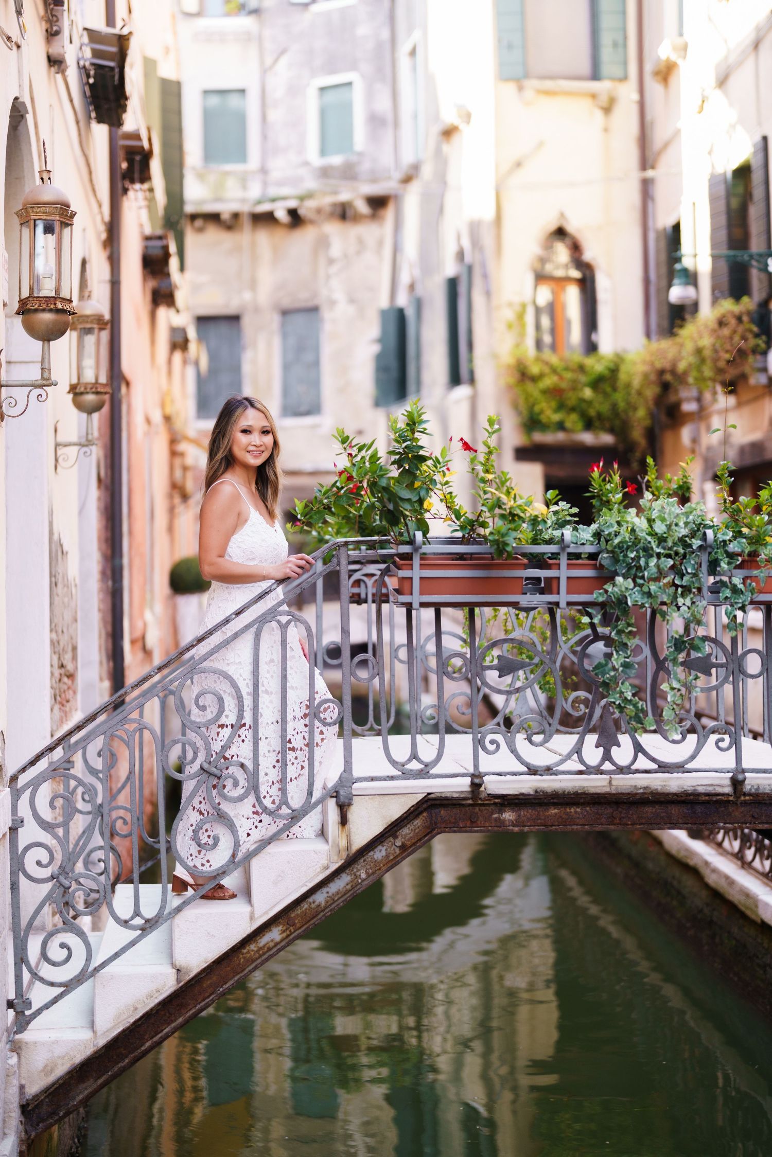 Young woman in white dress standing on ornate bridge over Venice canal surrounded by colorful flowers and historic buildings.