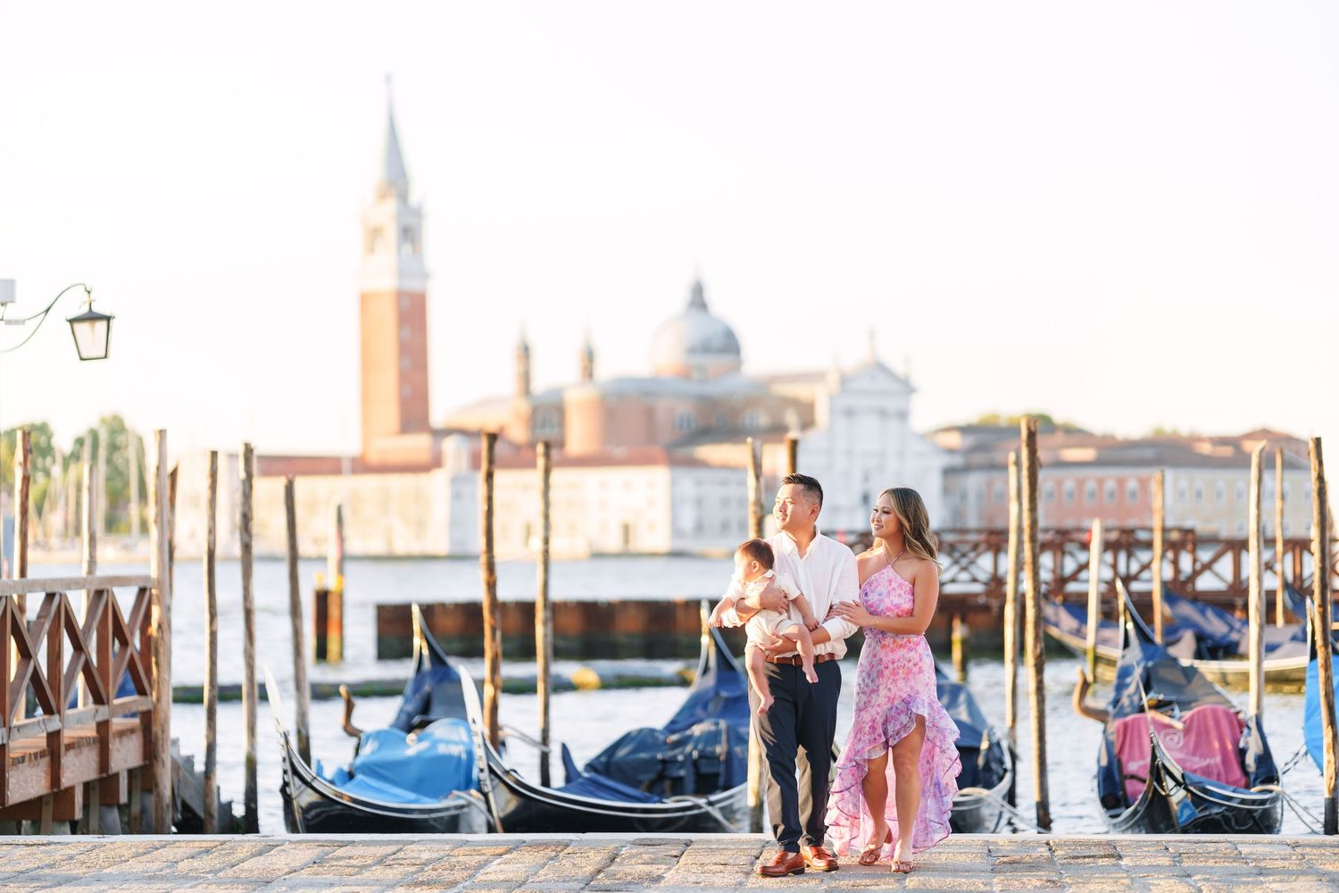 Couple standing on Venice dock with gondolas and San Giorgio Maggiore church in golden morning light.
