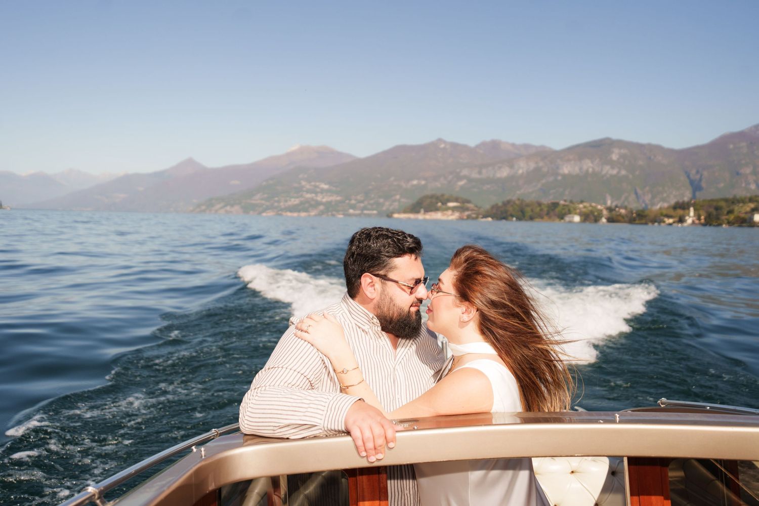 Couple sharing a romantic kiss on a wooden boat with stunning Lake Como mountains and blue water in the background.