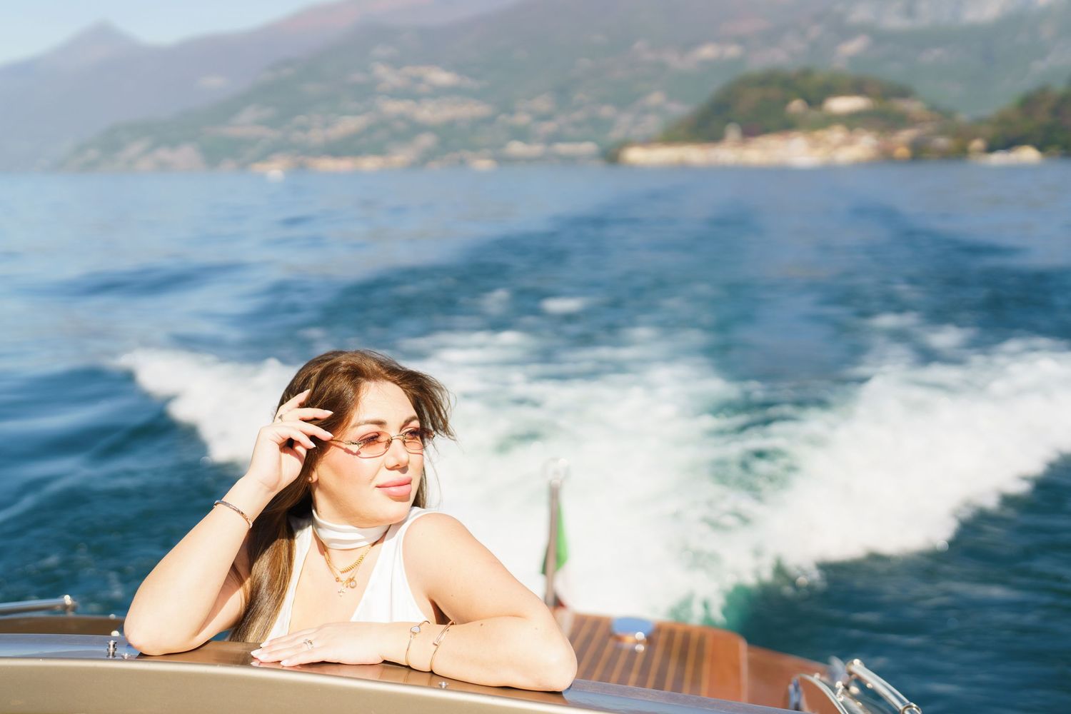 Woman in sunglasses relaxing on a luxury speedboat with scenic mountain lake views and blue water wake.