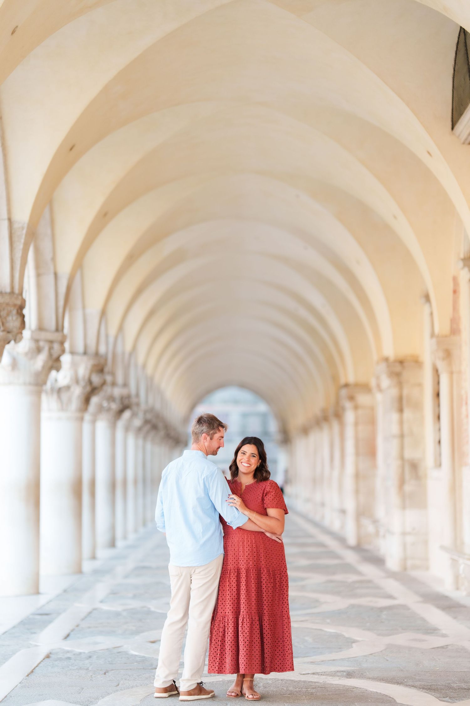 Couple embracing under elegant arched colonnade in Venice, woman in red dress, man in light blue shirt.