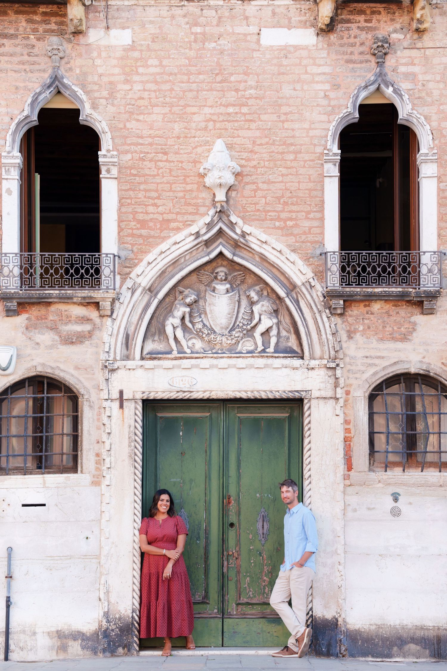 Couple posing by ornate Gothic green door of historic brick building with decorative arch in Venice, Italy.