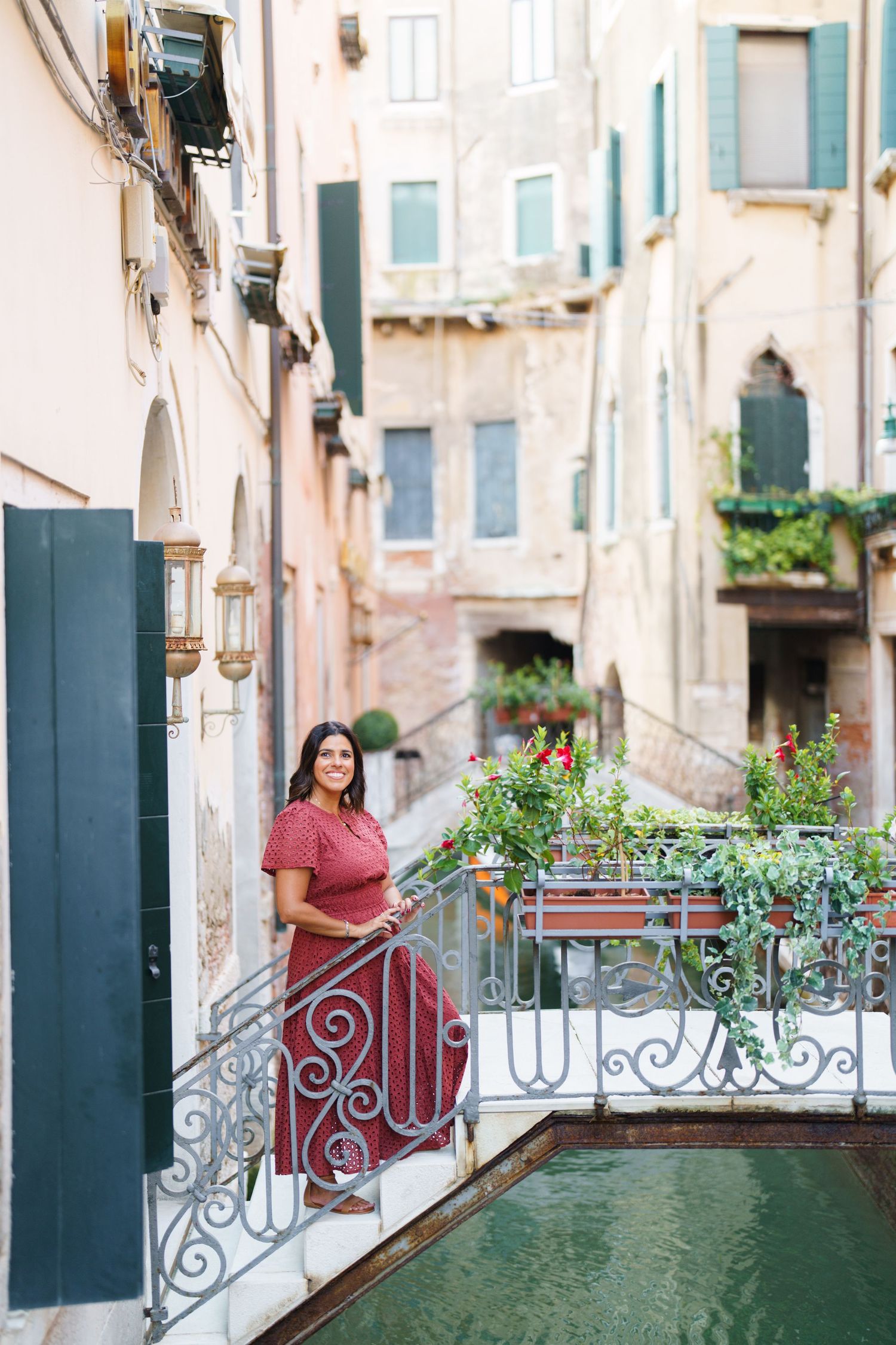 Woman in red dress standing on ornate bridge over Venice canal surrounded by colorful buildings and lush greenery.