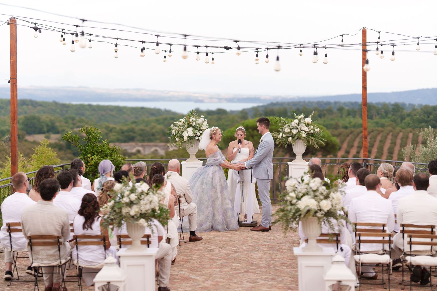 Outdoor vineyard wedding ceremony with white floral arrangements, string lights, and scenic rolling hills in the background.