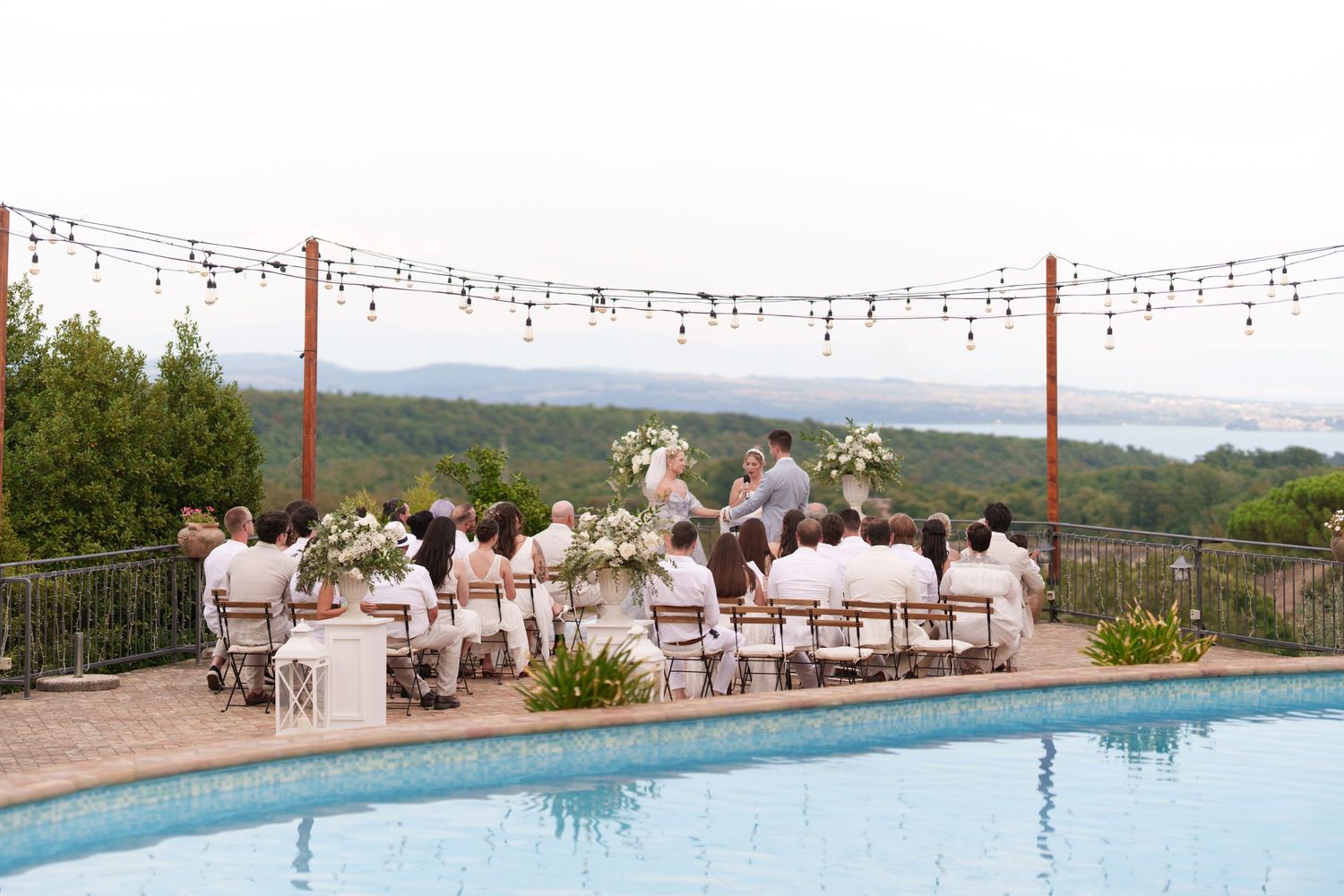 Outdoor wedding ceremony by a pool with string lights, floral arrangements, and scenic valley views.