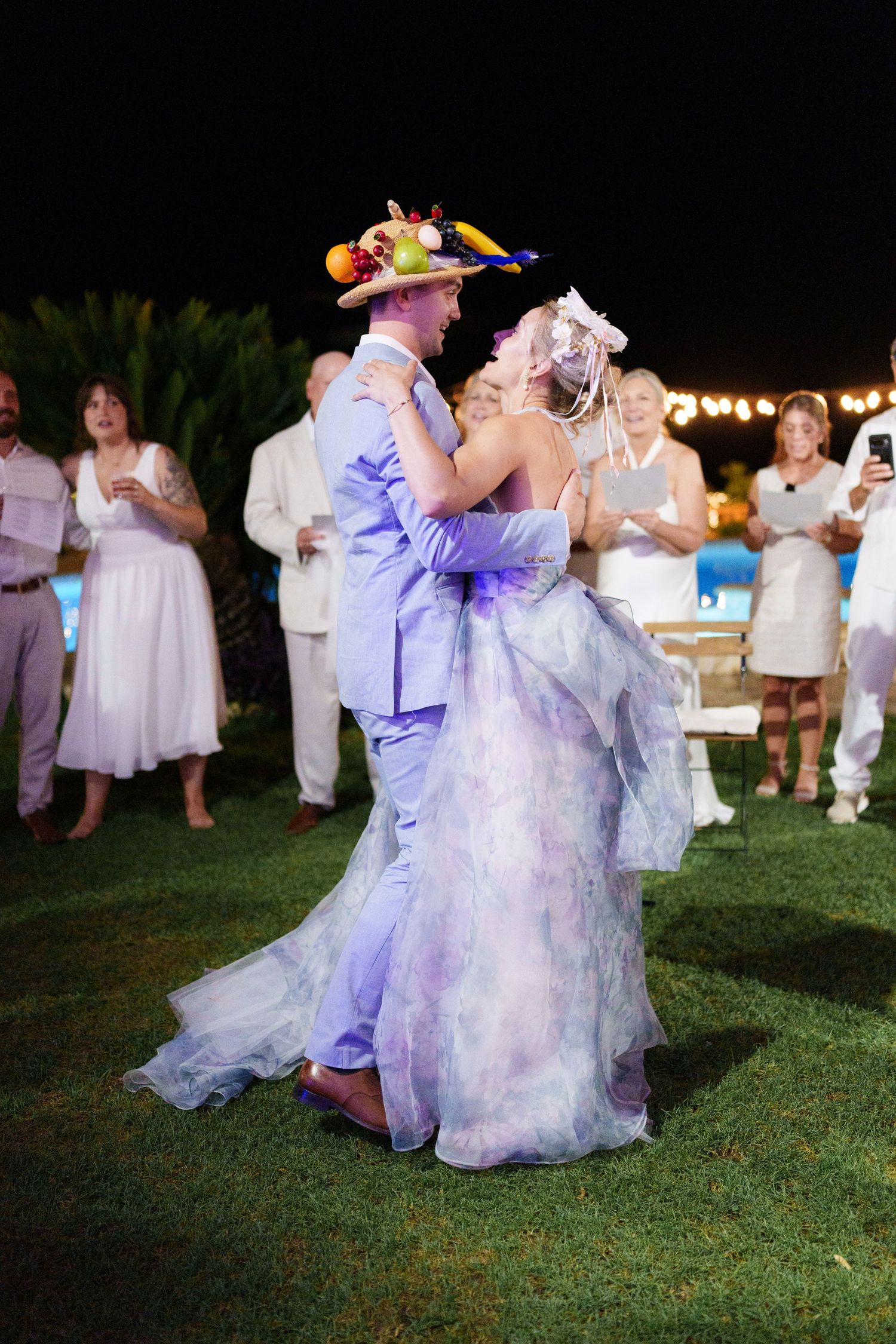 Couple sharing first dance at outdoor wedding reception, bride in purple gown, groom in light blue suit with fruit hat.