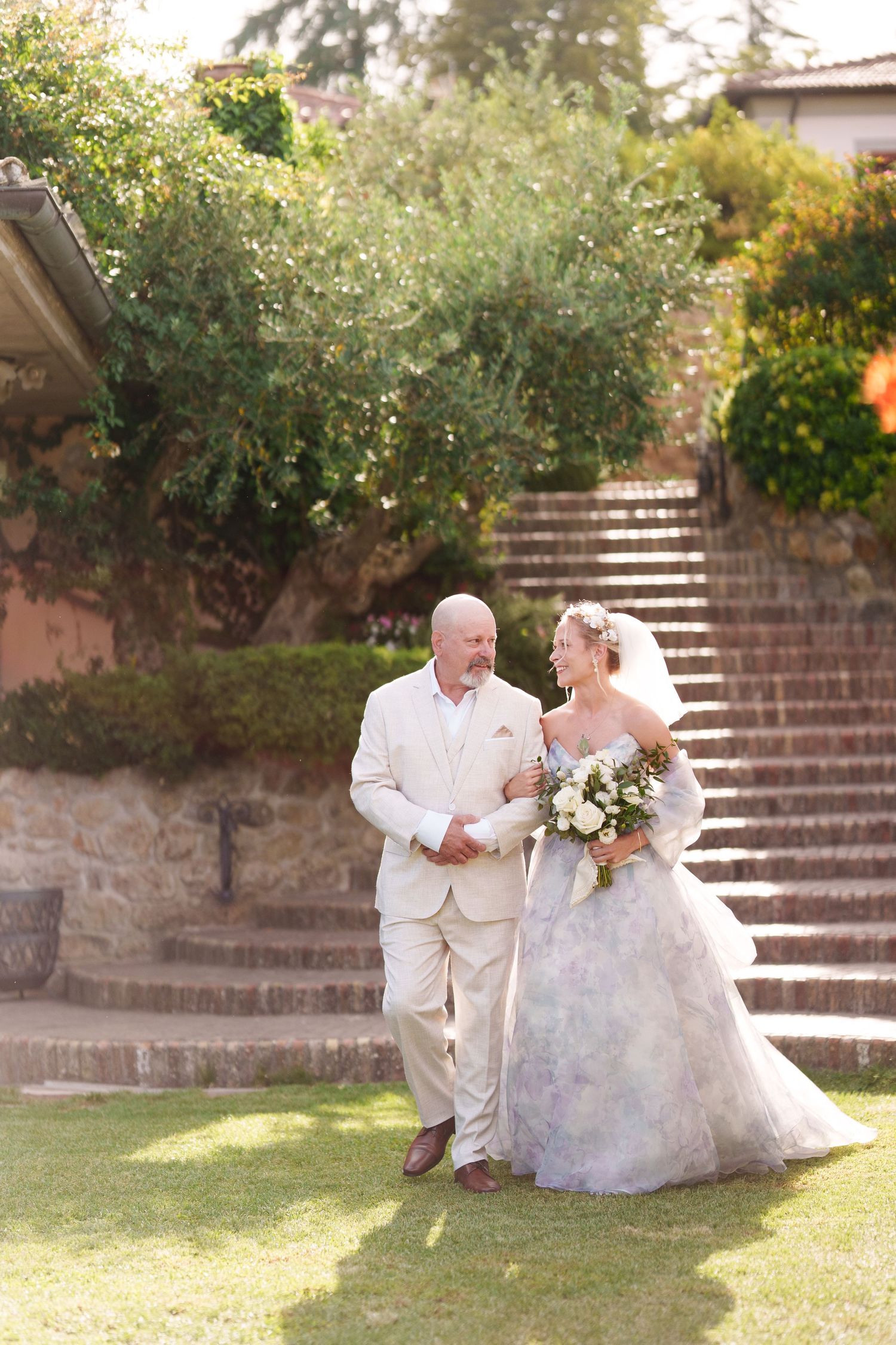 Bride in floral gown holding bouquet walks with groom in beige suit on garden lawn with stone steps backdrop.