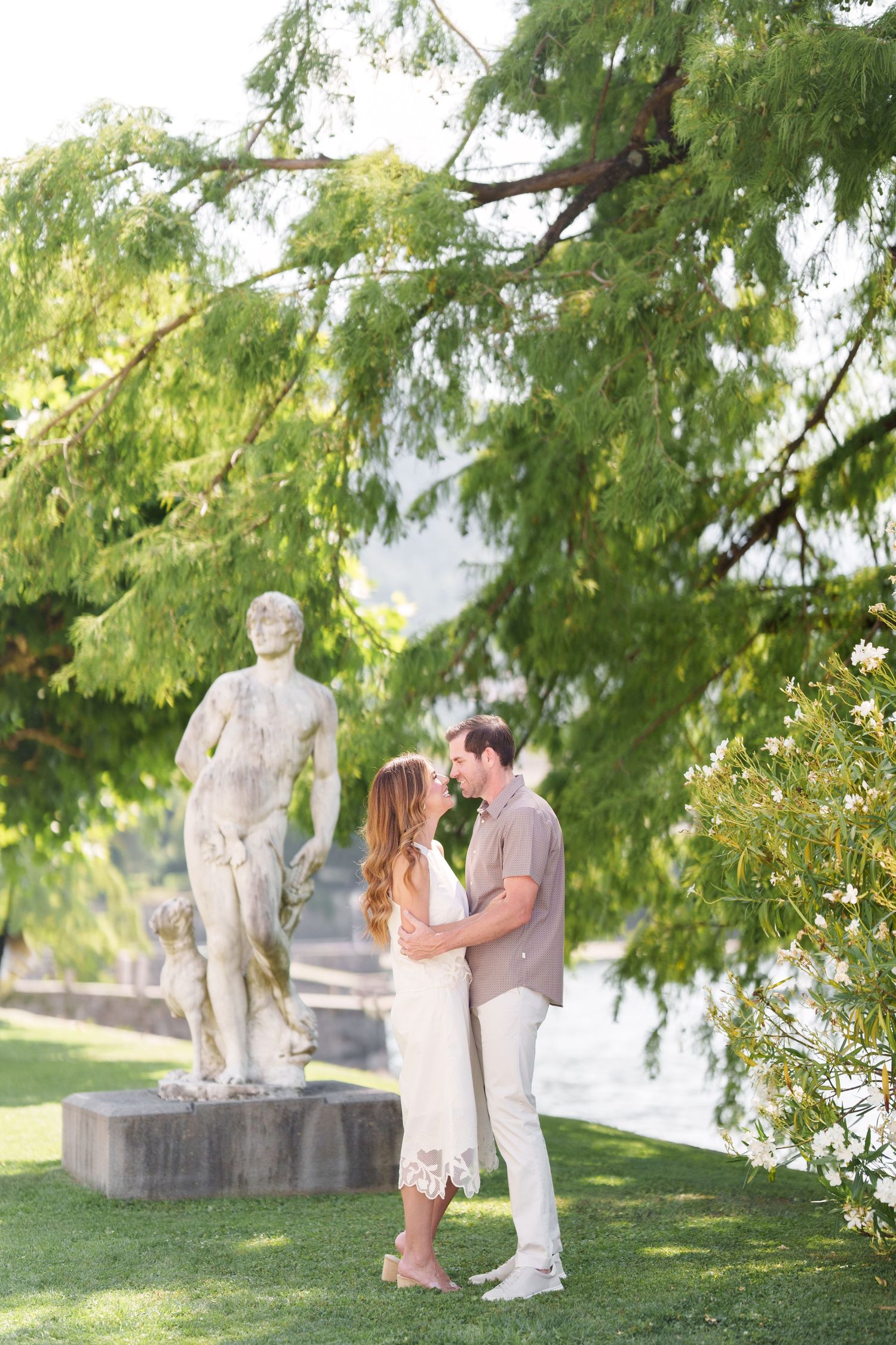 Couple sharing a romantic kiss in a lush garden with a classical statue and green trees in the background.