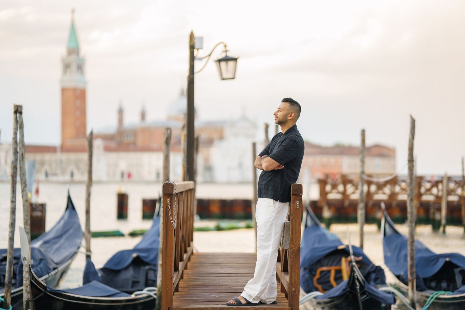 Man in white pants and navy shirt stands on Venice gondola dock with San Giorgio Maggiore church in background.