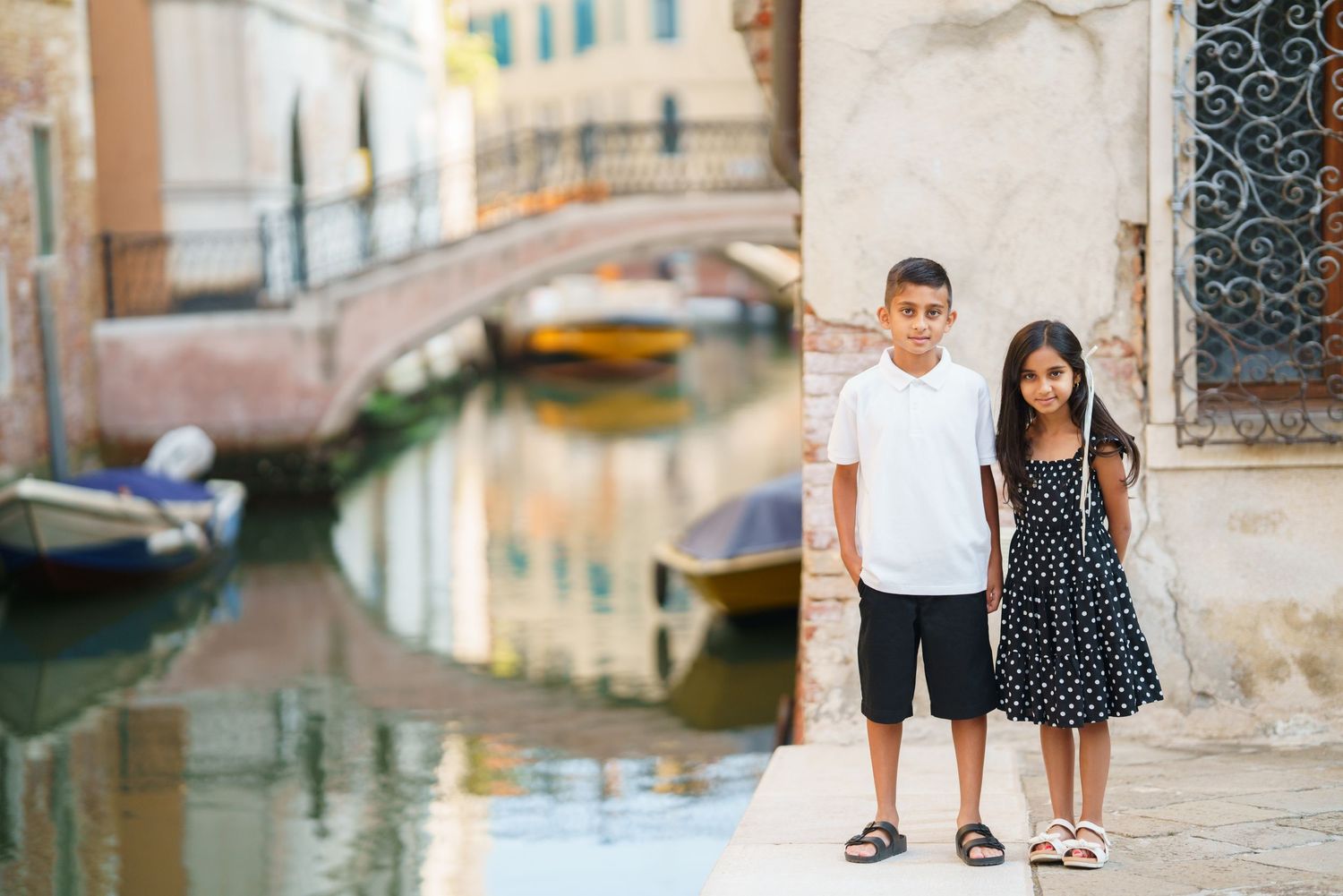 Two children pose along a Venice canal with historic bridge and colorful boats in the background.