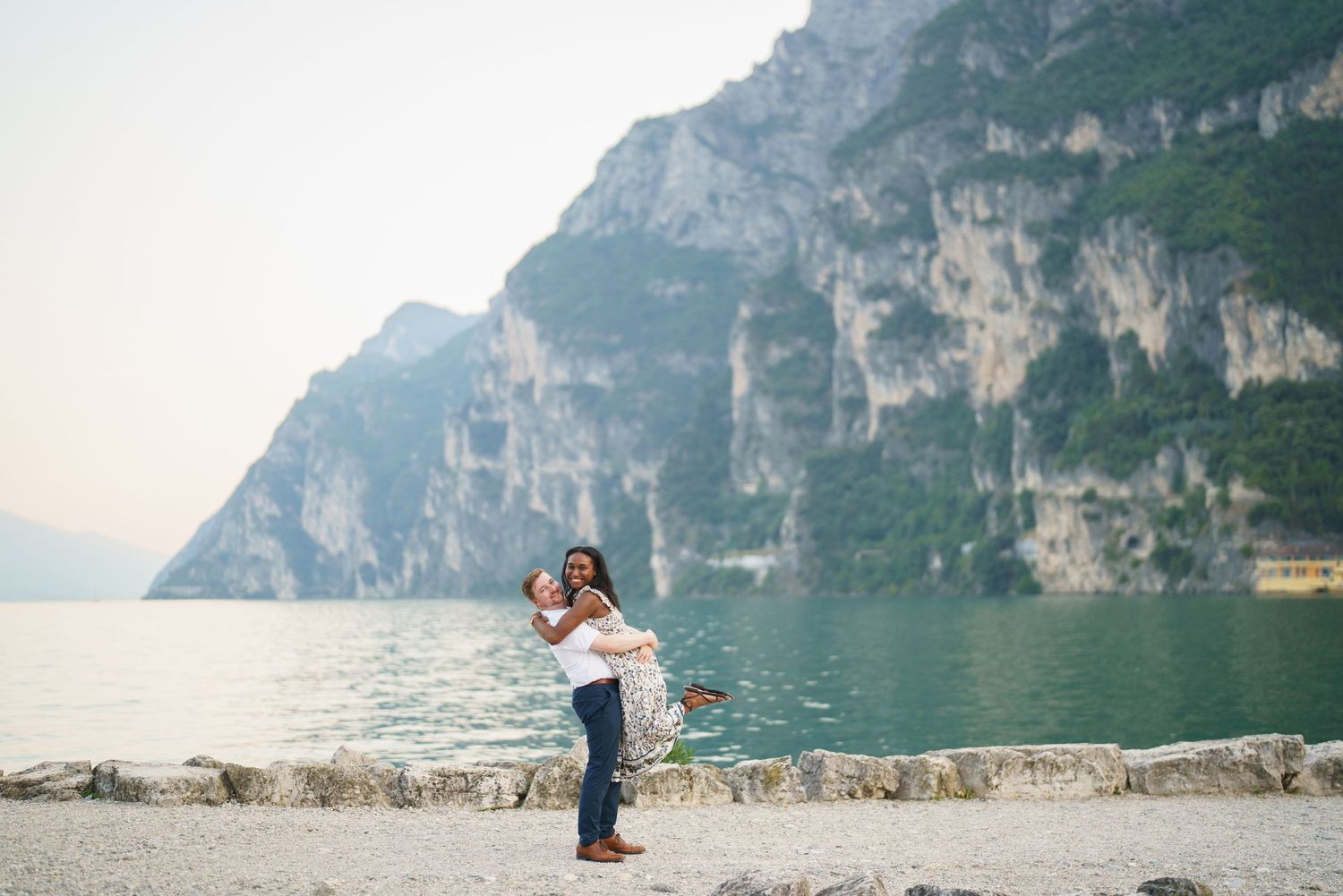 Joyful interracial couple embracing and spinning by a stunning alpine lake with mountains.