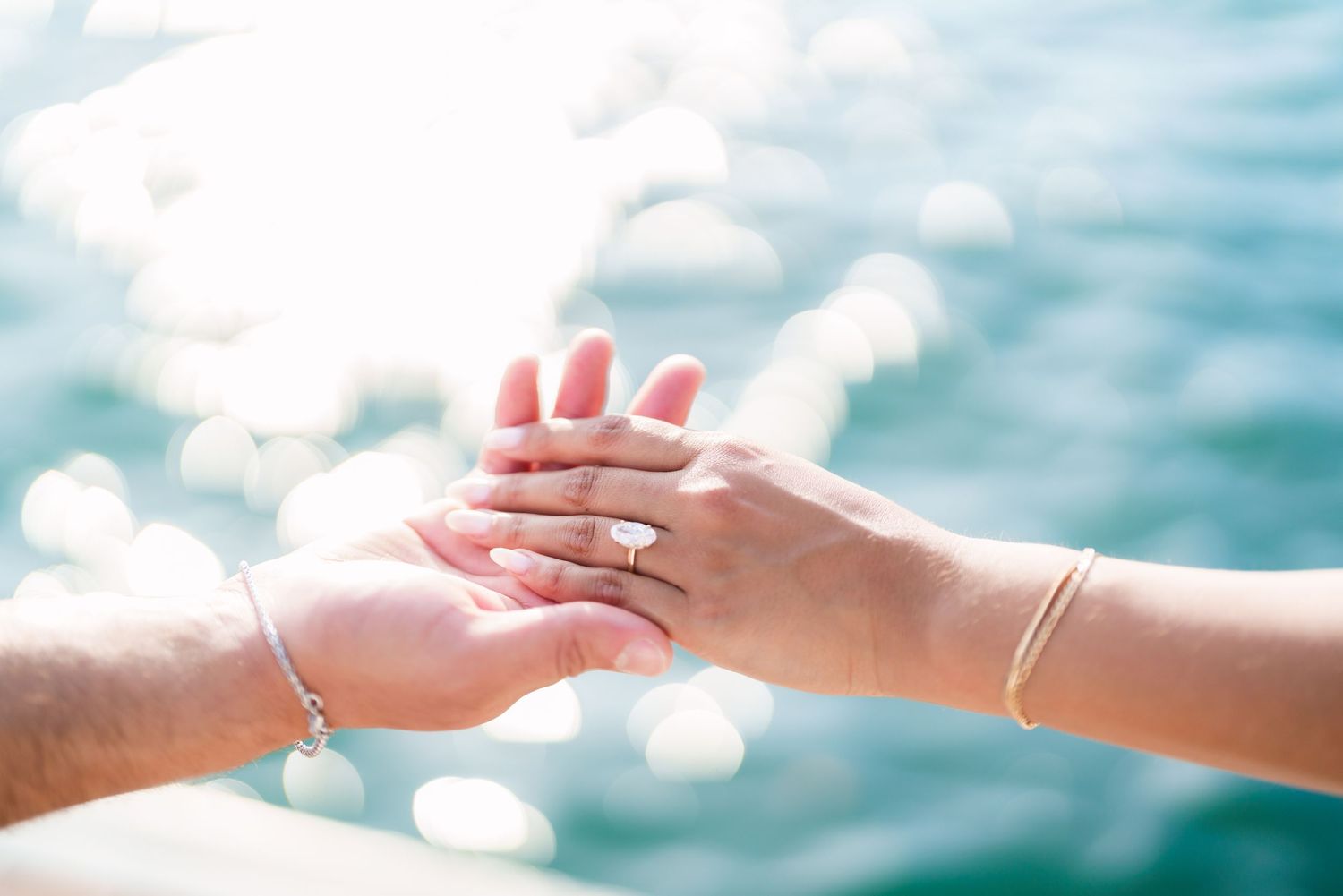 Two hands gently touching over sparkling blue water, one adorned with an engagement ring and bracelet.