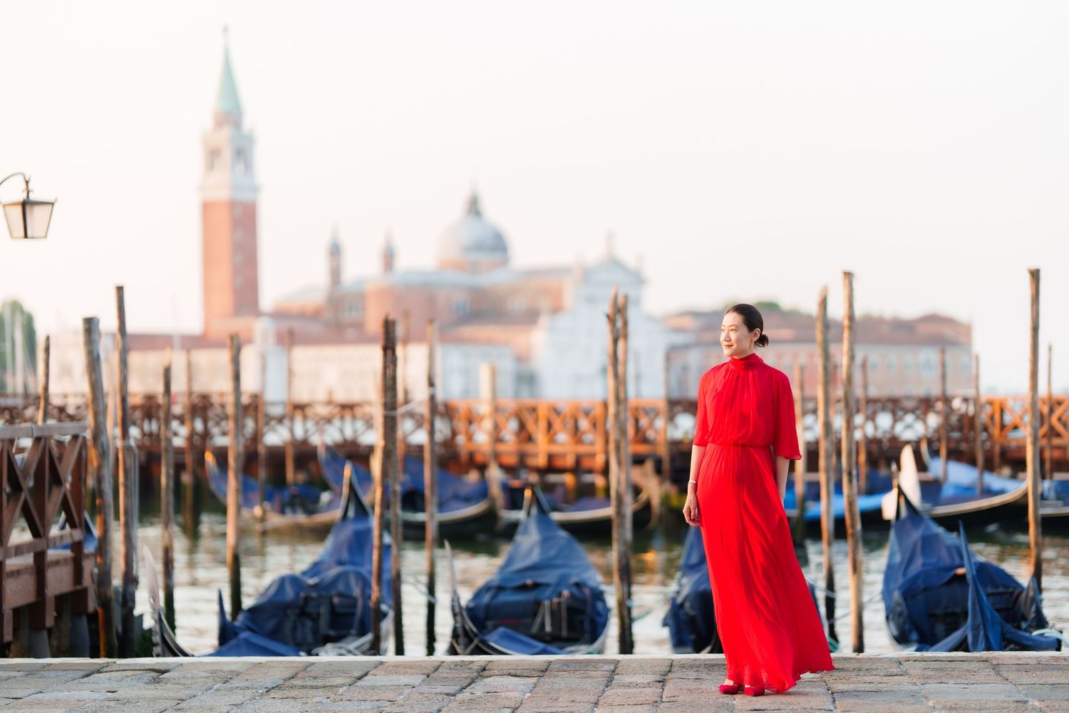 Woman in red dress standing by Venice gondolas with San Giorgio Maggiore church in background.
