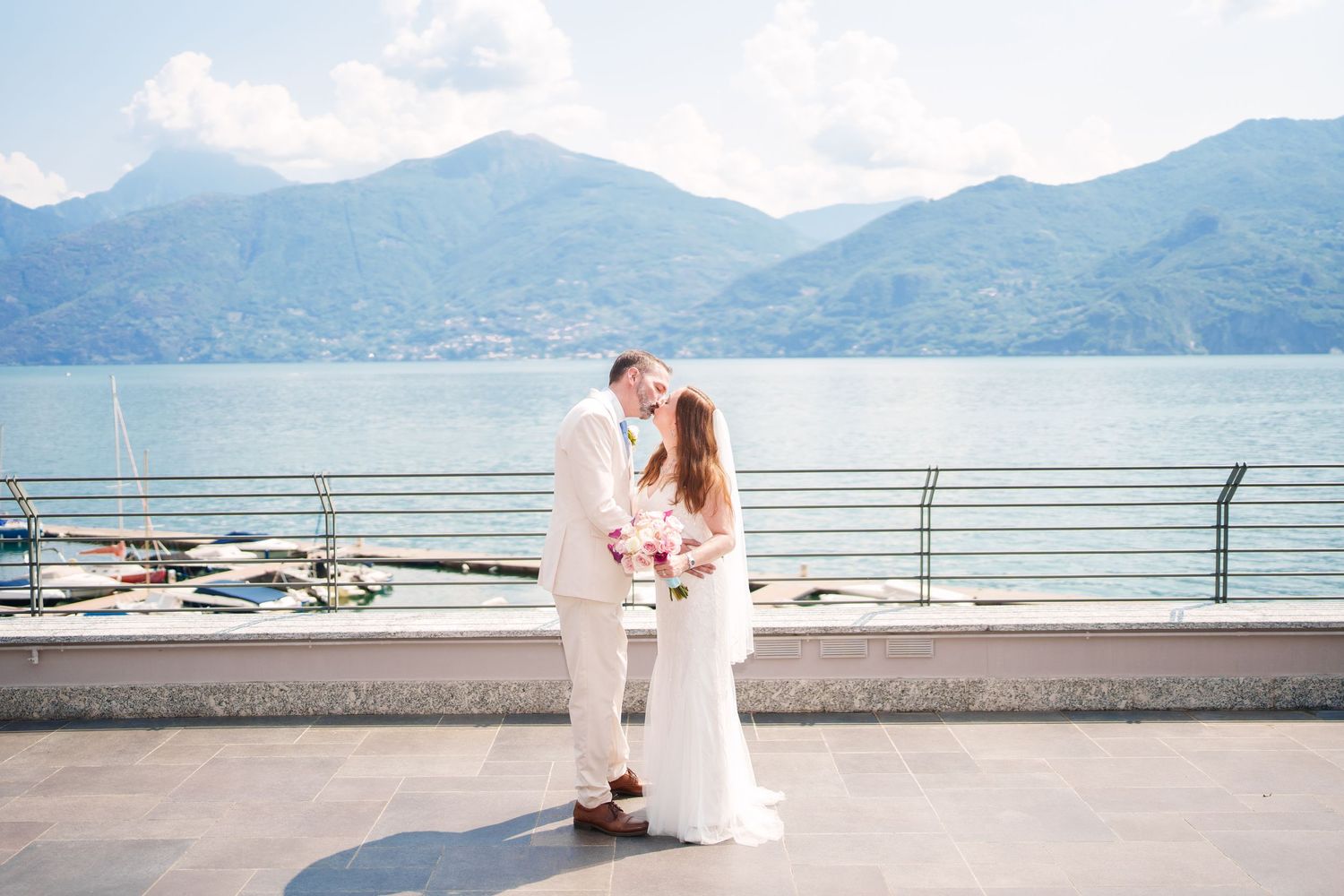 Bride and groom share a romantic kiss on a lakeside terrace with stunning mountain views in the background.