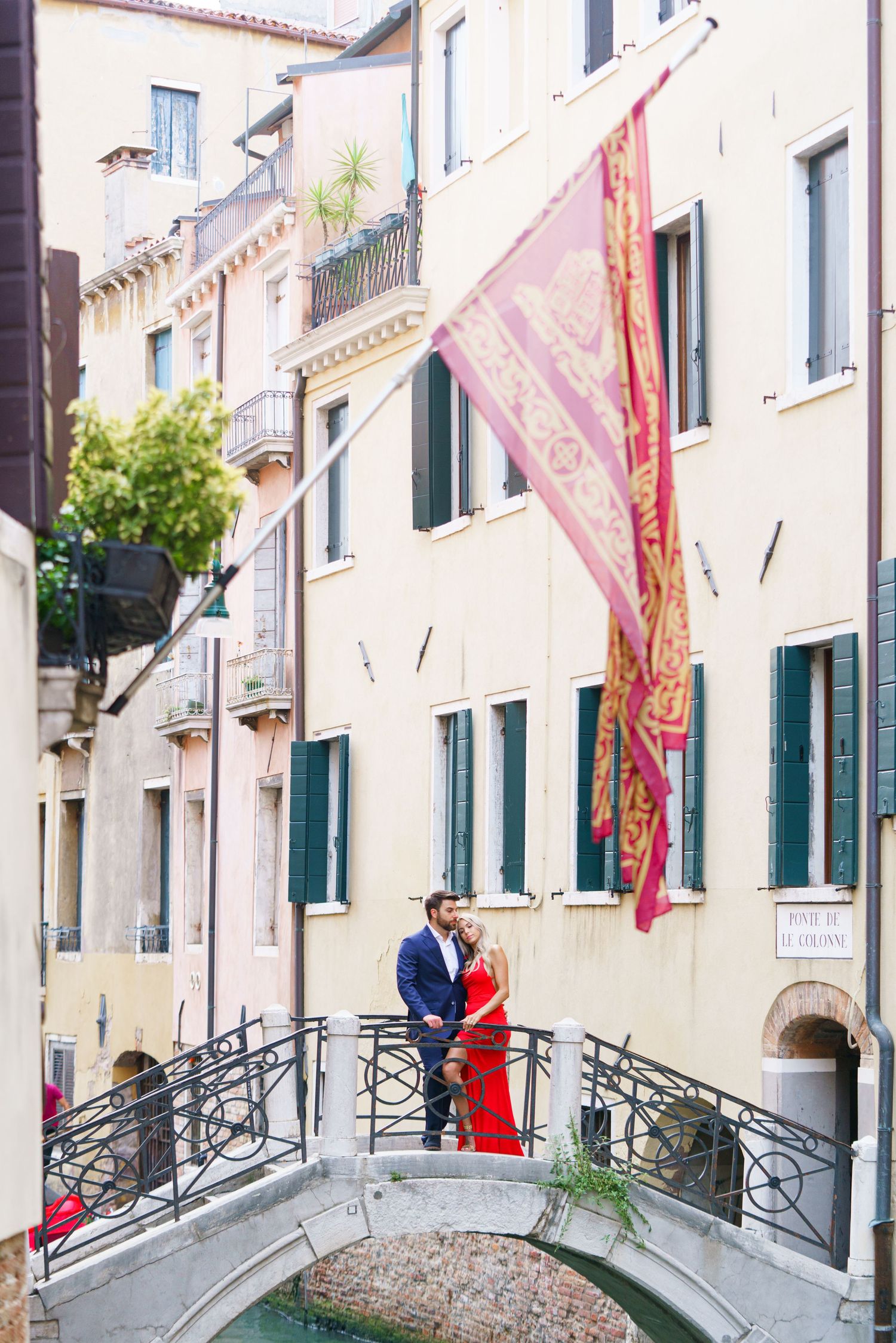 Two people in formal attire stand on a romantic Venetian bridge with traditional architecture and flags in the background.