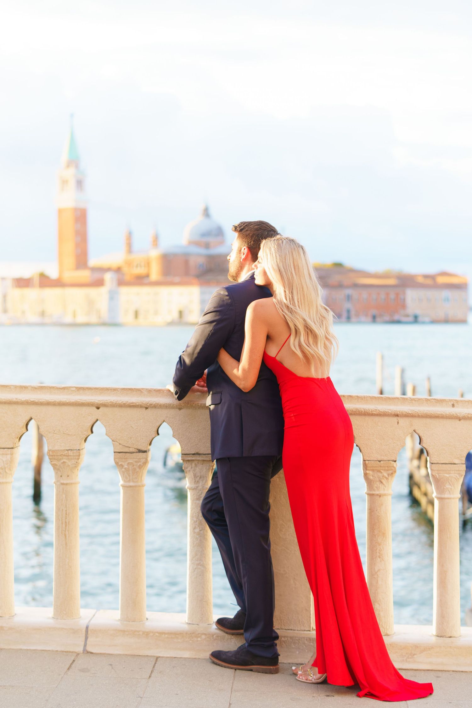 Couple in formal attire poses along a waterfront balcony in Venice with the iconic skyline in the background.