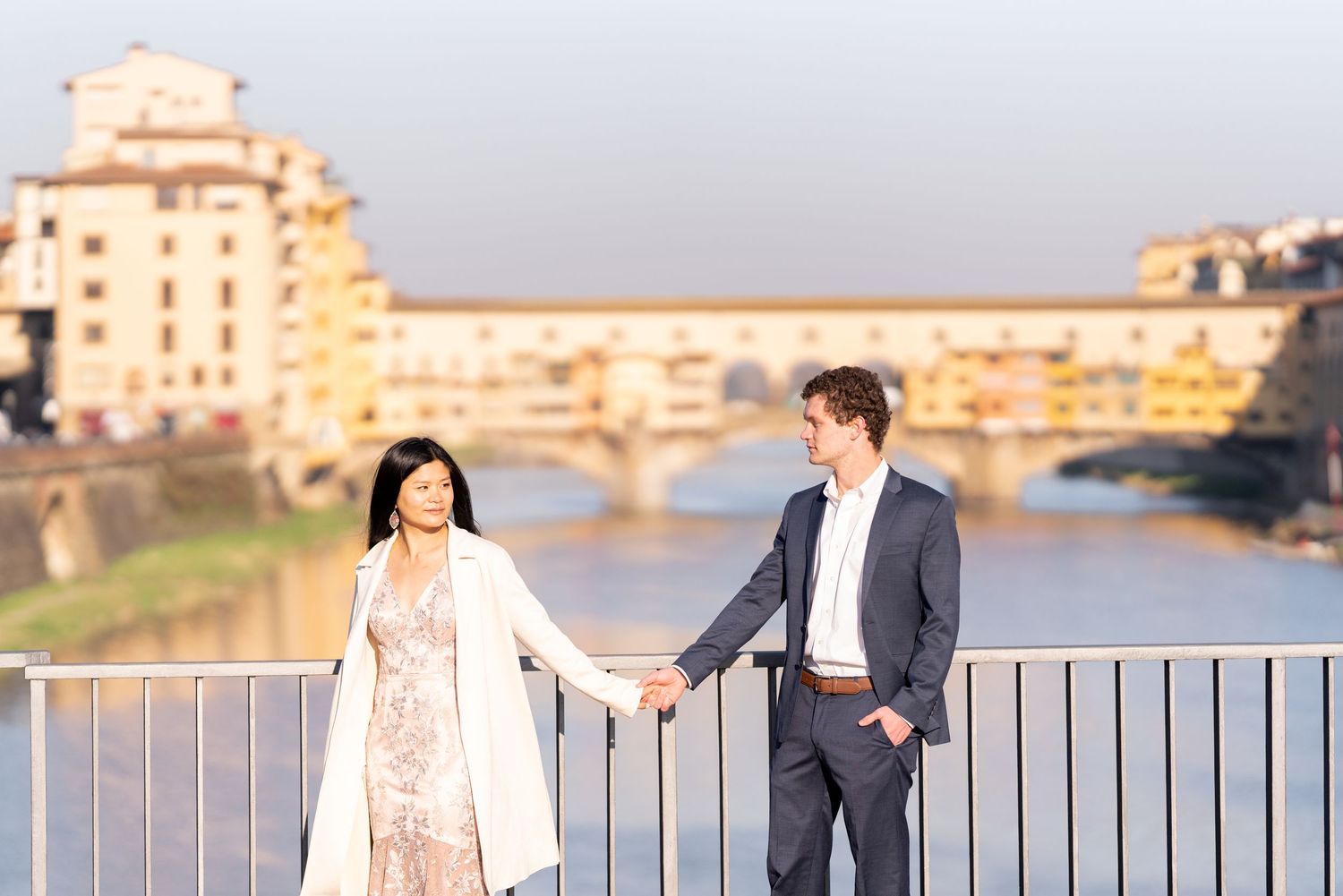 A couple poses together along a railing overlooking Florence's Ponte Vecchio bridge during a golden sunset.