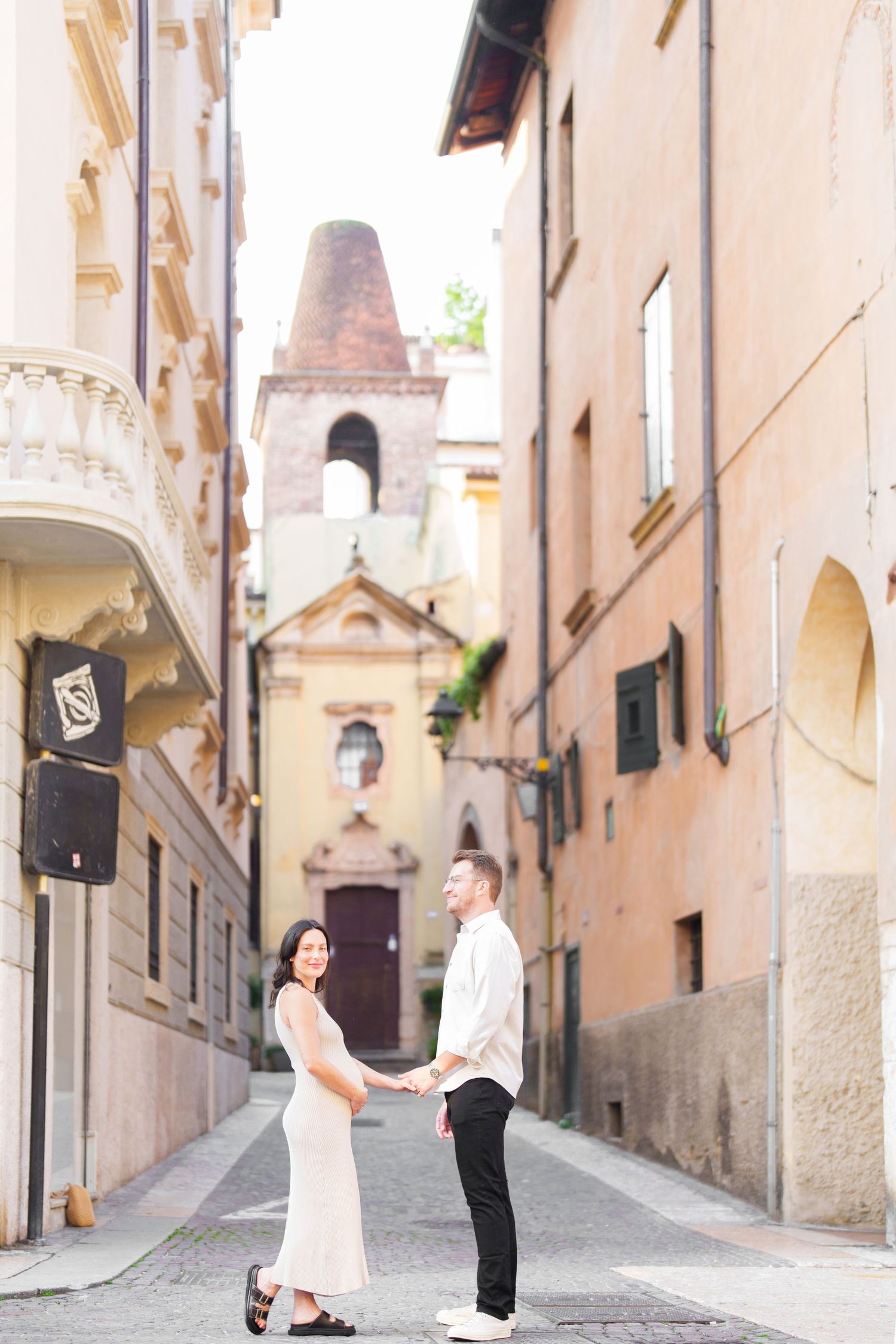 A romantic couple walks through charming narrow cobblestone streets in a European old town with peach-colored buildings.