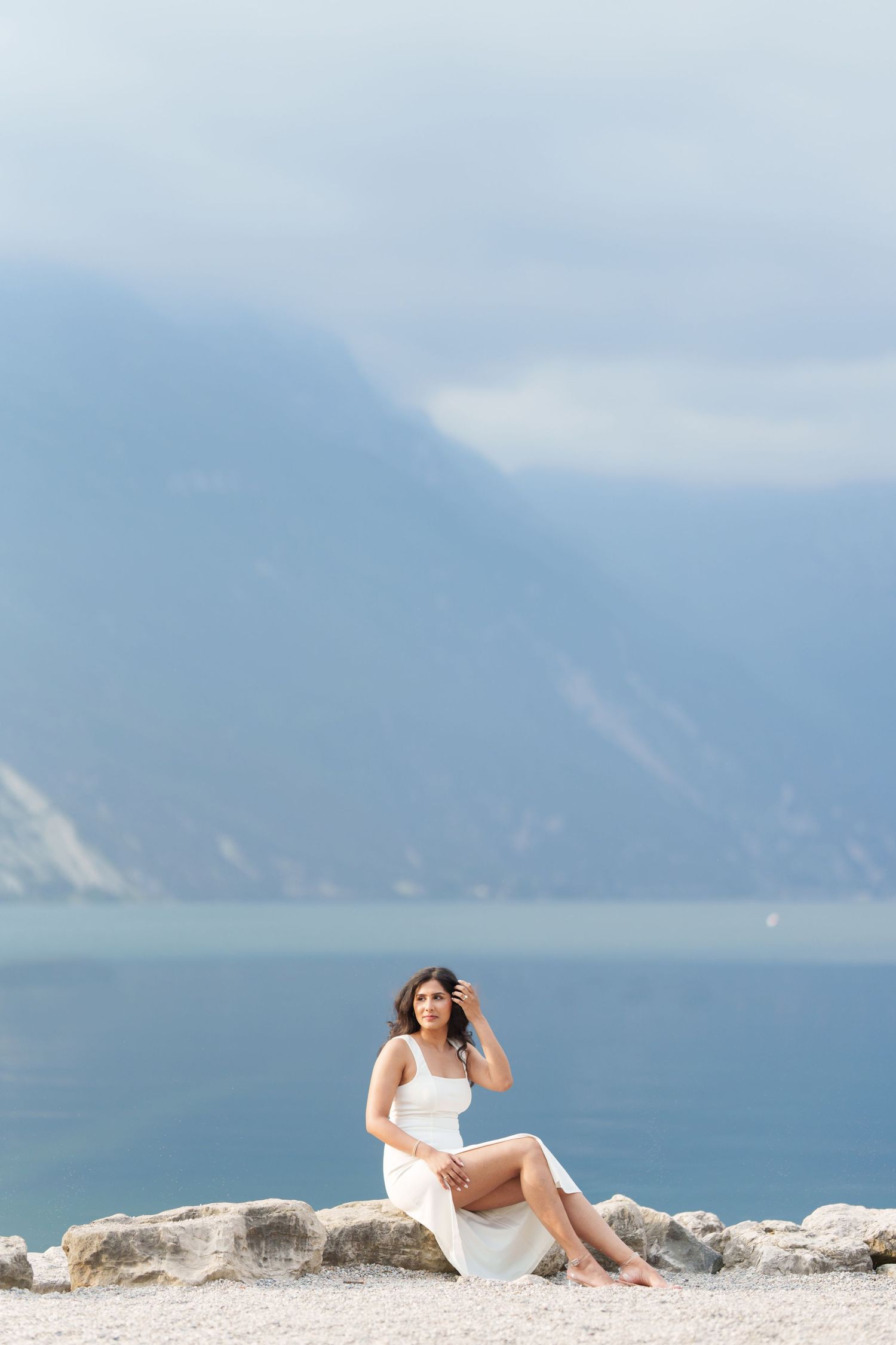 A woman in a white dress sits gracefully on coastal rocks with misty mountains and ocean in the background.