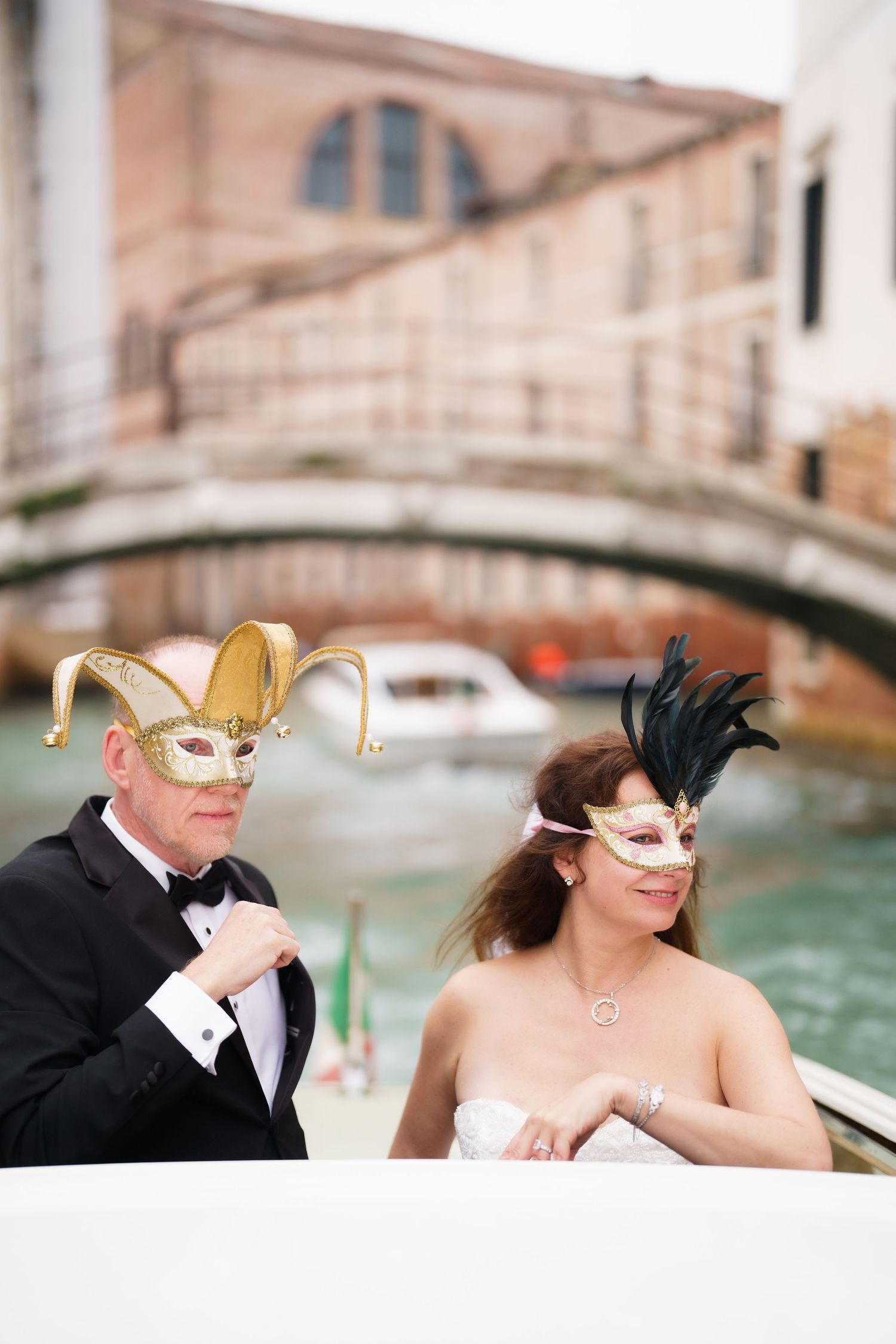 A couple wearing gold carnival masks pose together on a gondola in Venice with historic bridges and buildings in background.
