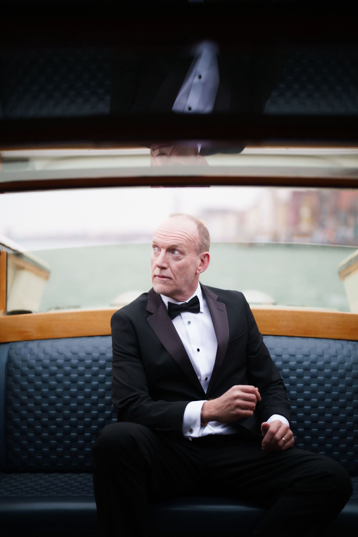 A person in a tuxedo sits on a dark leather couch in a dimly lit room, looking to the side pensively.