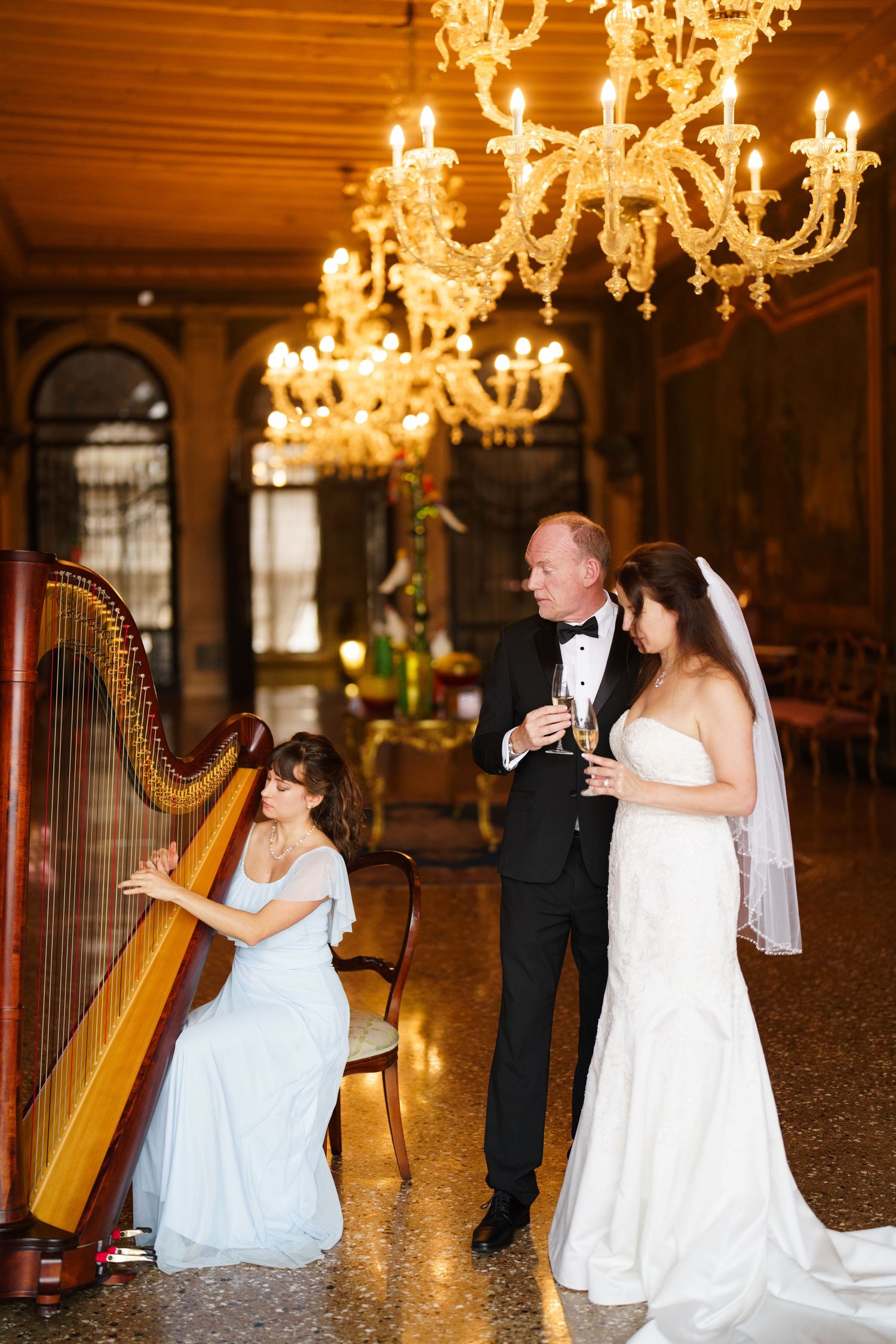 Romantic wedding couple dances together under ornate crystal chandeliers in an elegant ballroom.