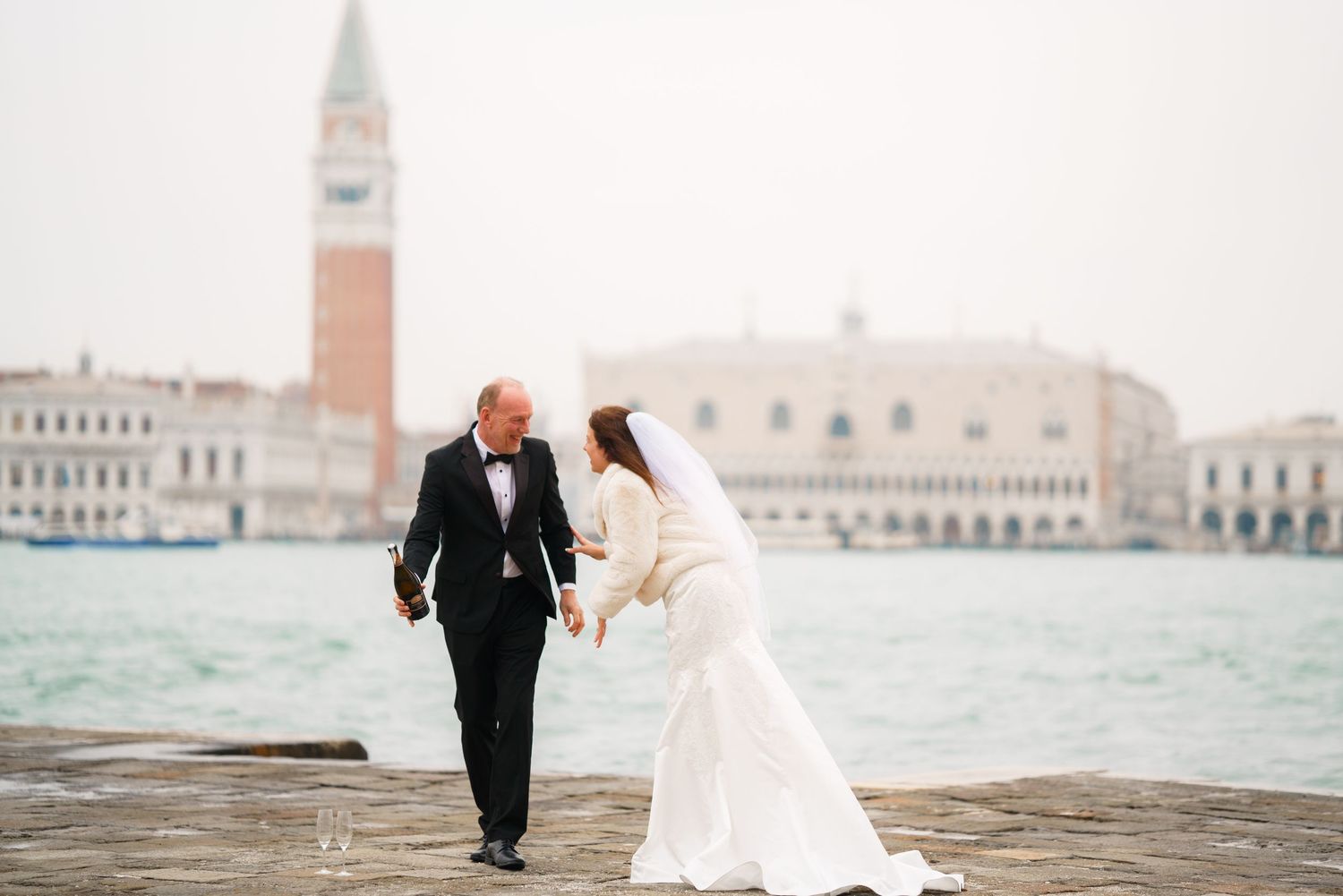 A romantic wedding couple walks along Venice's iconic waterfront with historic buildings and gondolas in view.