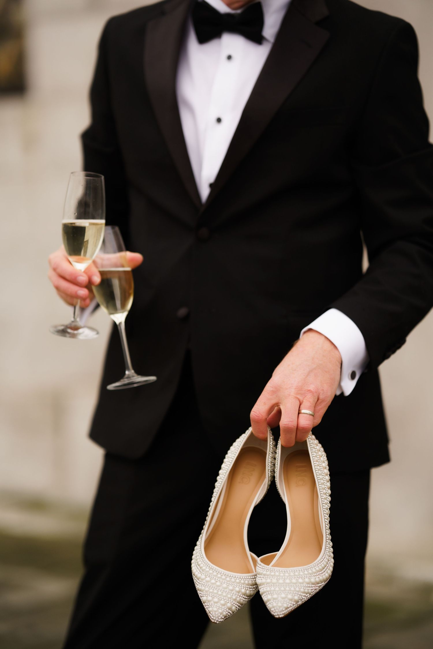 Elegant white pearl-studded bridal flats held by someone in a black tuxedo with champagne glass.