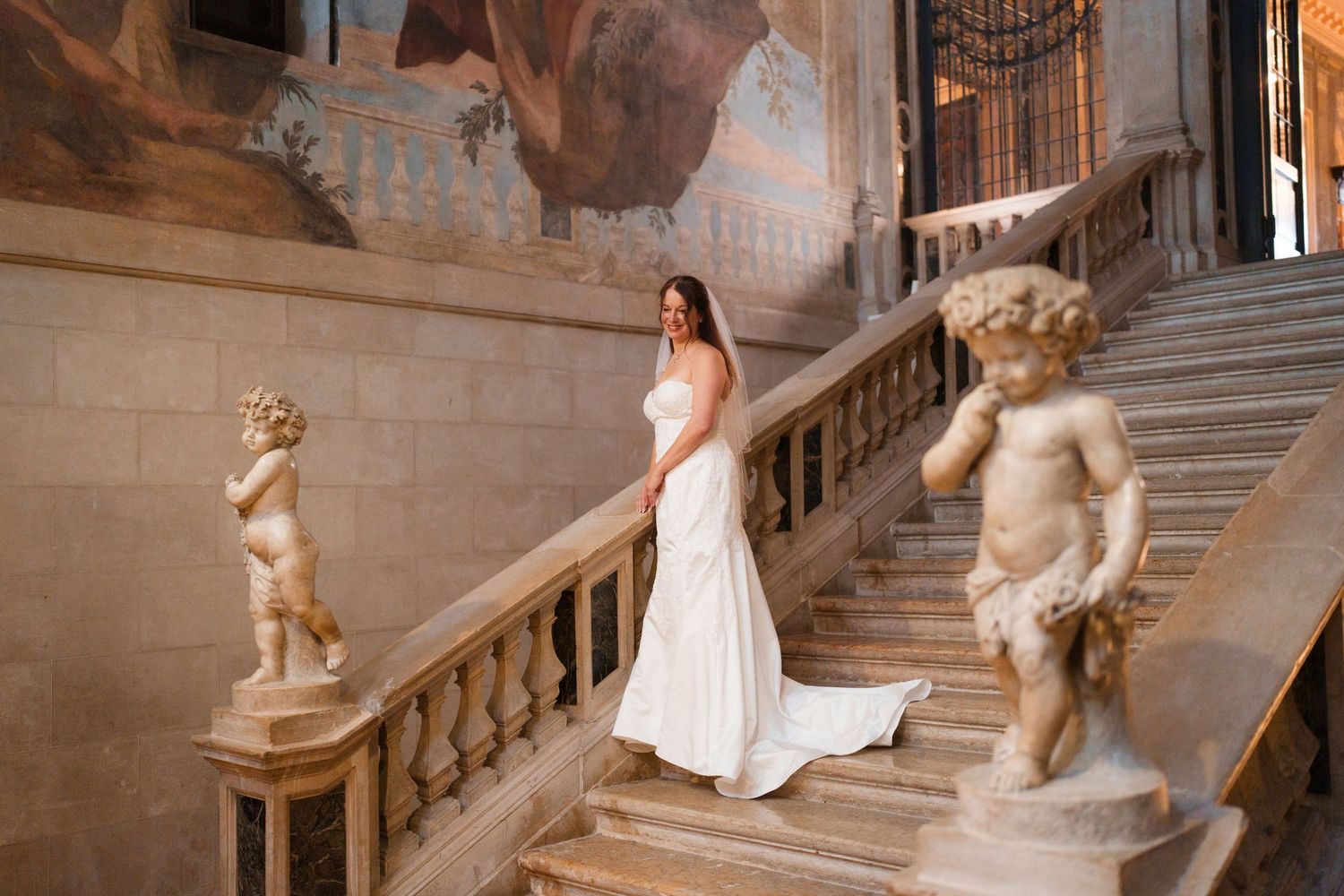 A bride in a flowing white gown poses elegantly on a grand marble staircase adorned with classical statues.