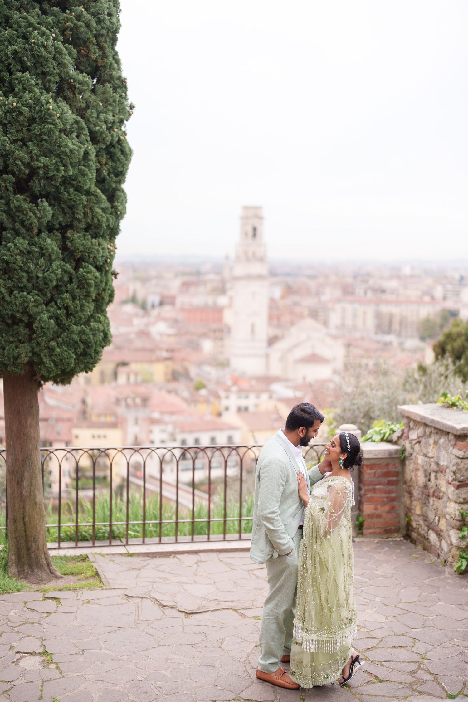 A couple shares romantic moments on a stone terrace overlooking a scenic European cityscape with terracotta rooftops.