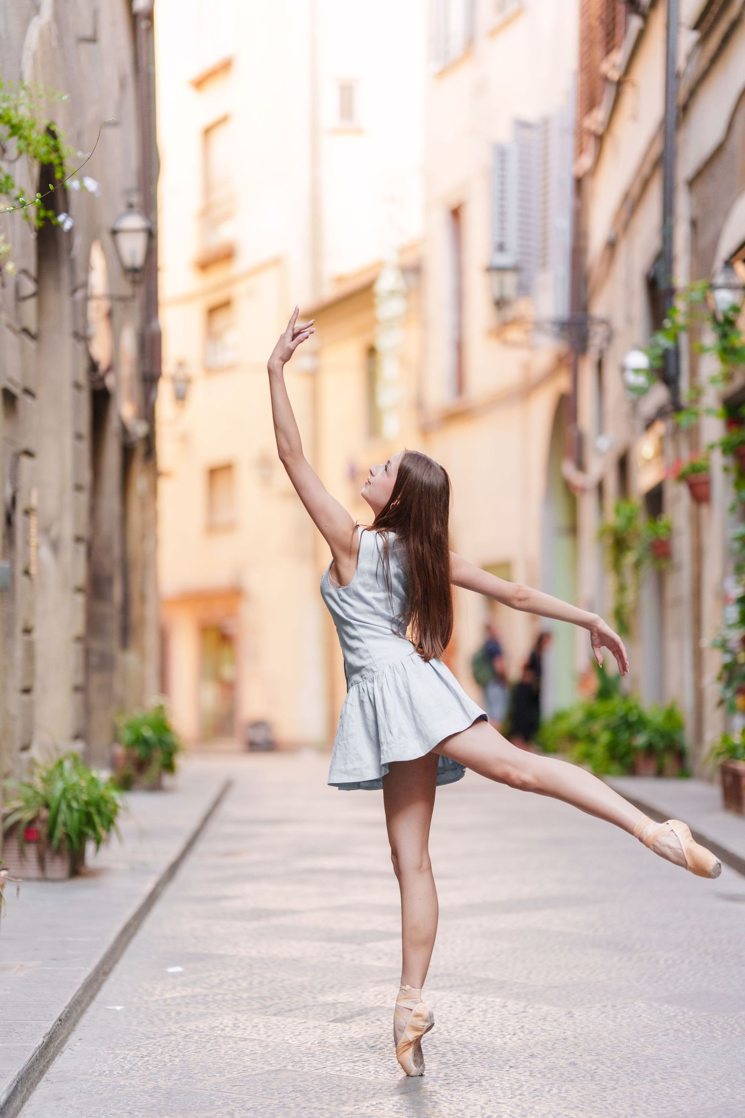 A ballet dancer in a light blue dress performs an elegant pose on pointe shoes in a charming European alleyway.