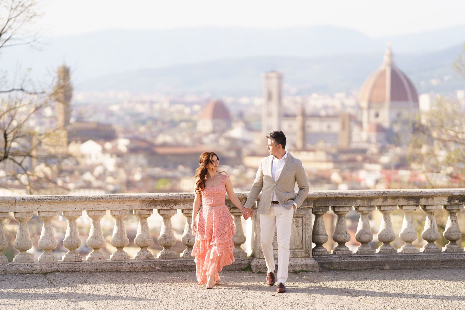 Romantic couple in elegant attire walking along Piazzale Michelangelo terrace overlooking Florence's Duomo cathedral.