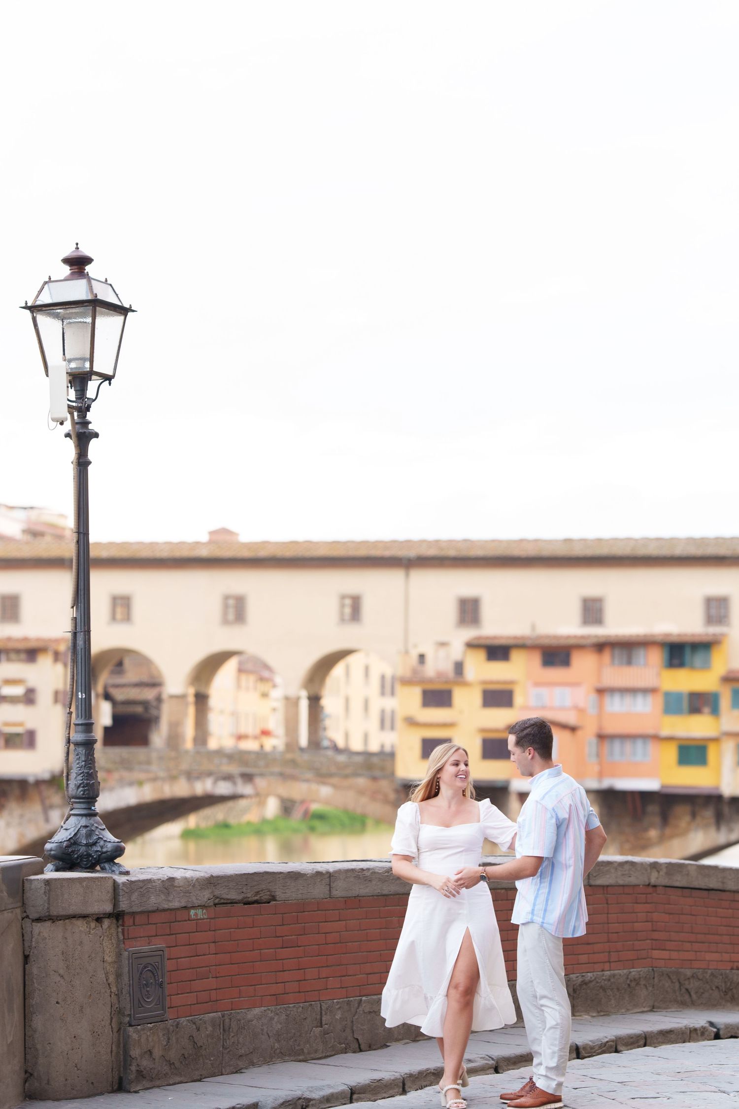 A couple in white outfits poses romantically along a historic brick wall in Florence, Italy at sunset.