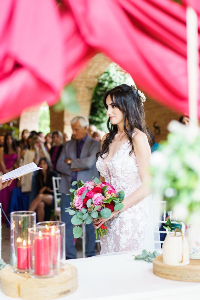 Bride holds pink and green bouquet during wedding ceremony with red drapery and candle decorations.