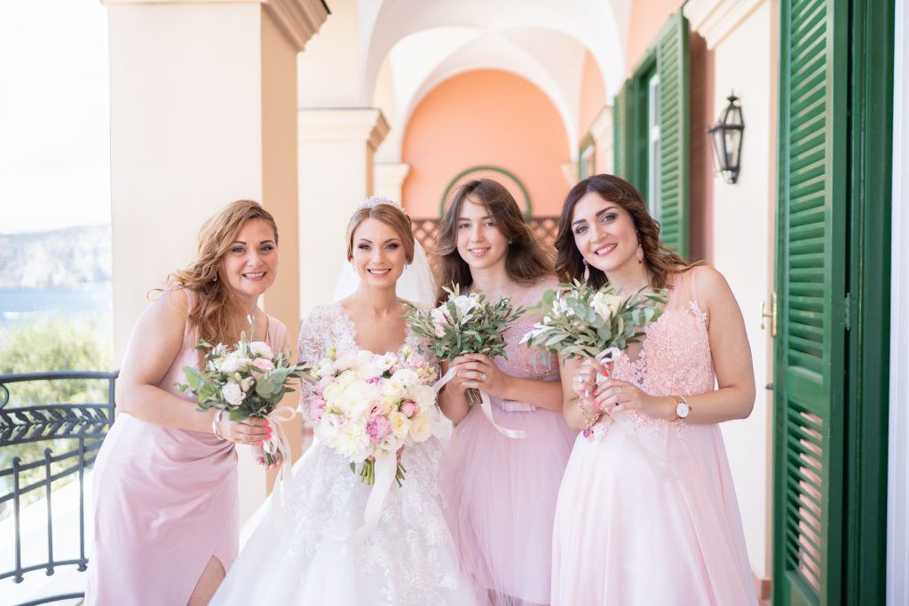 Bridesmaids in soft pink dresses pose together on an elegant terrace with white columns and green shutters.