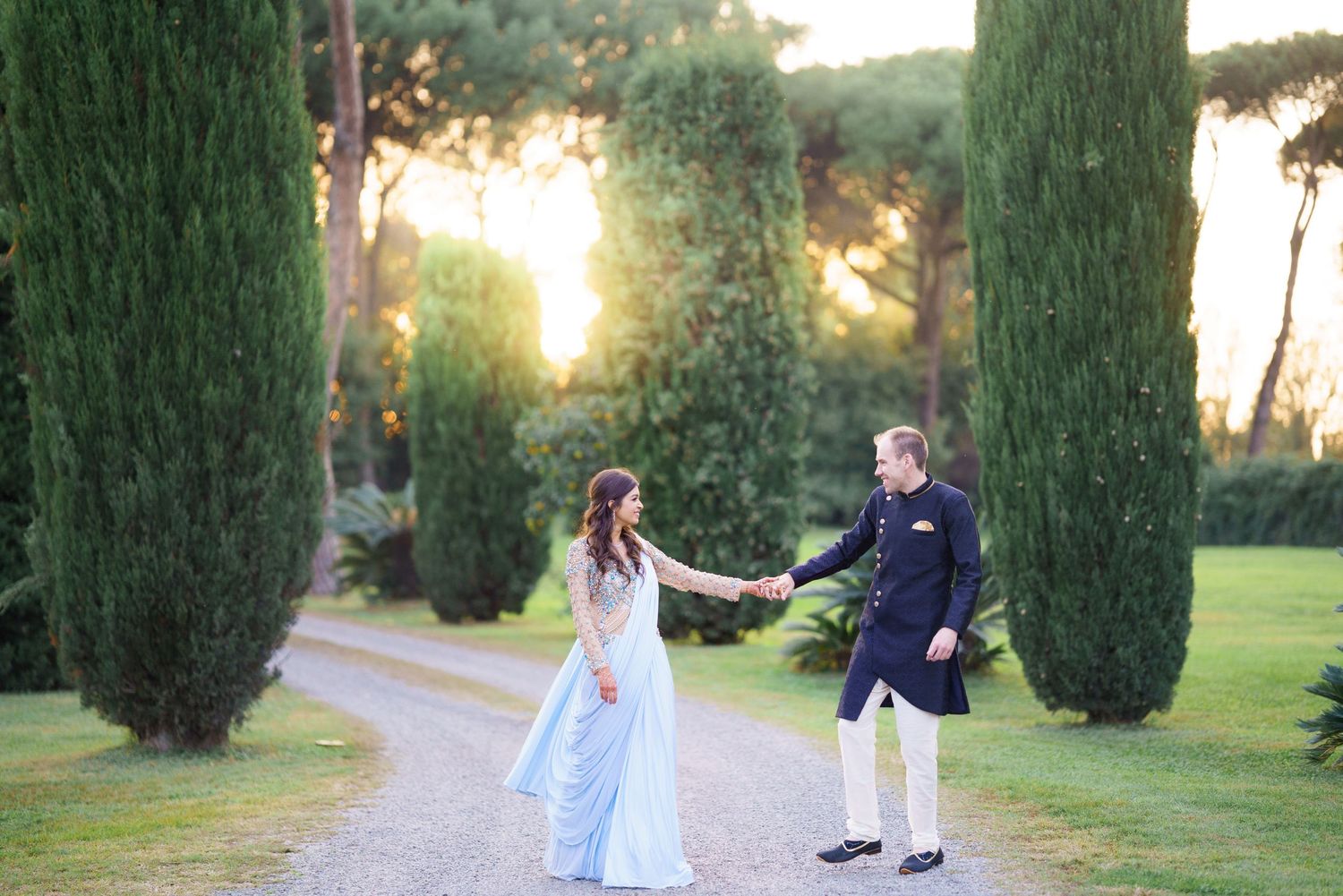Romantic couple poses in a garden setting with tall cypress trees during golden hour sunset.