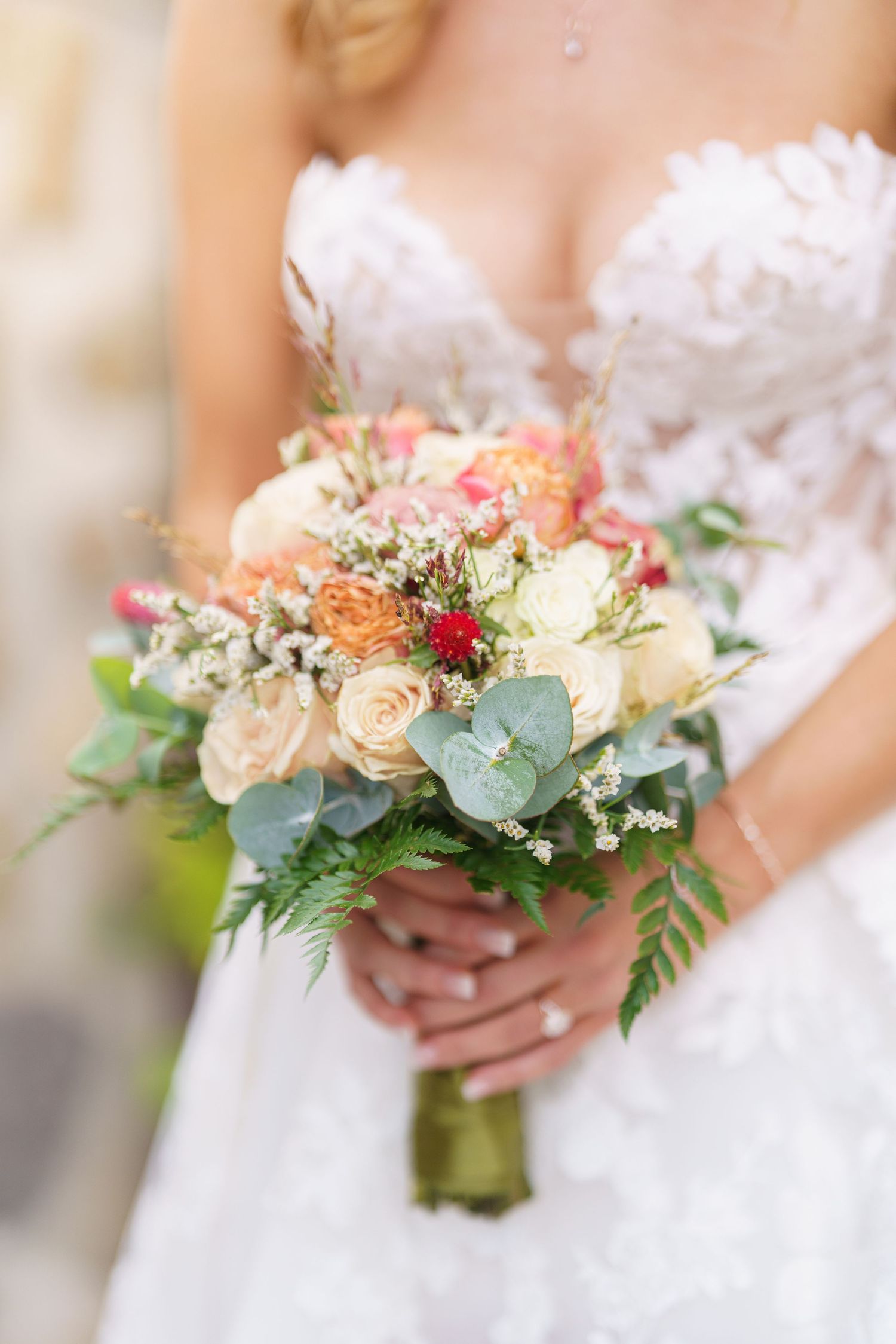 Wedding bouquet featuring peach roses, eucalyptus, and red accents wrapped in white ribbon.