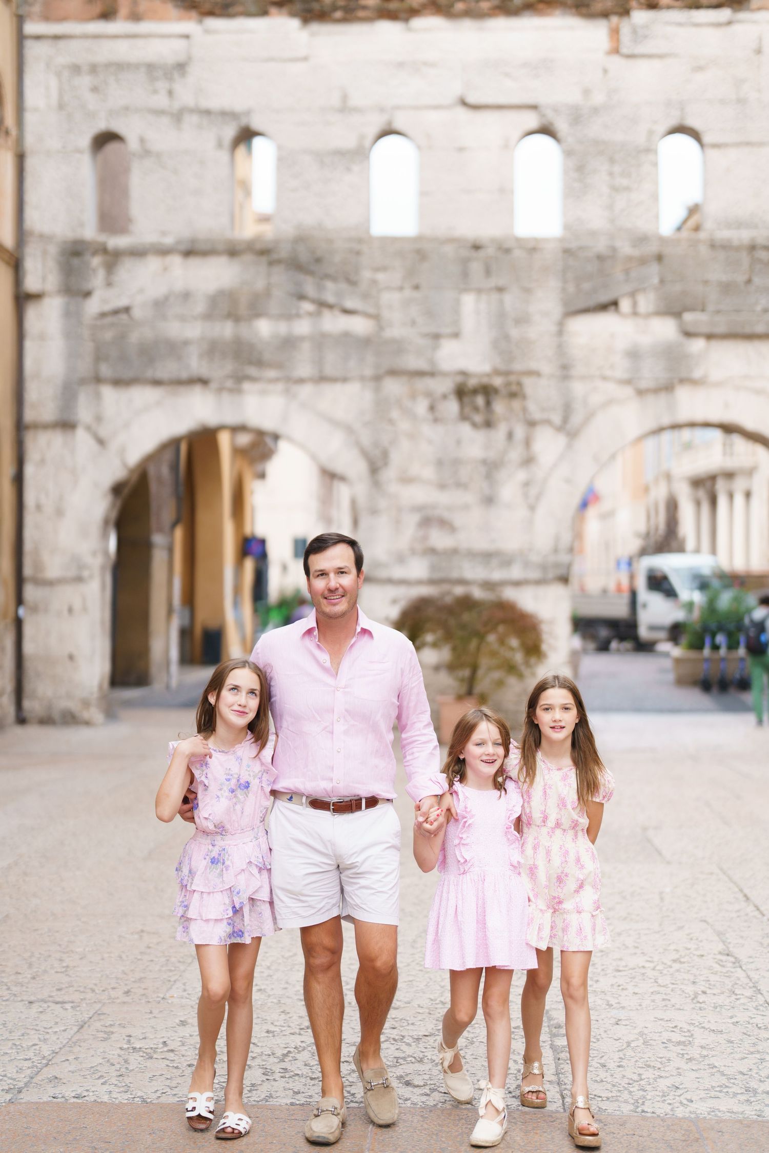 Father and three daughters in pink outfits posing in front of ancient Roman arch in historic European city.