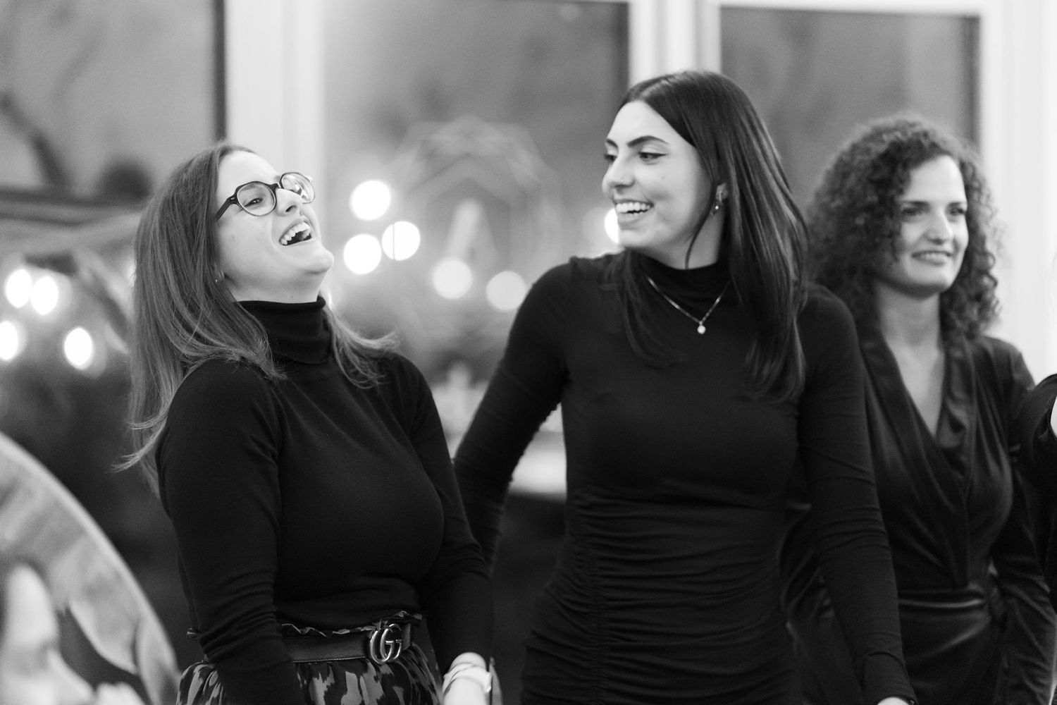 Three women in black outfits laughing together at an indoor event in a black and white photo.