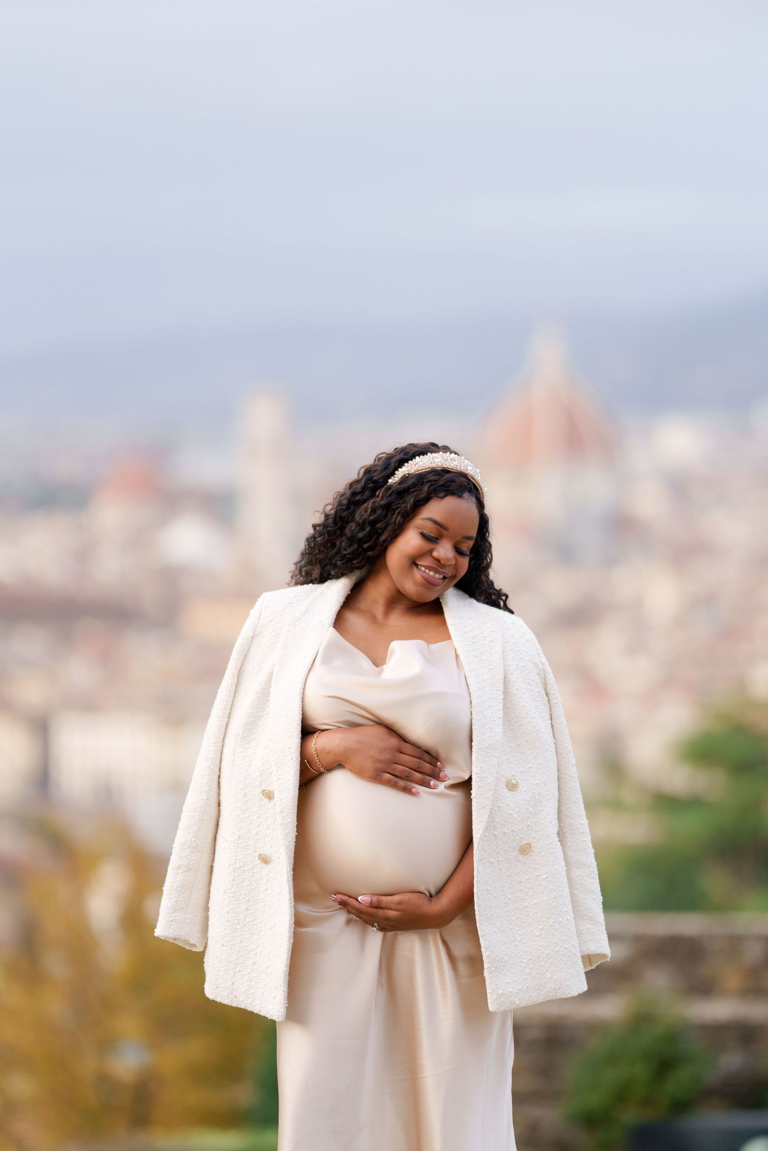 Smiling pregnant woman in cream dress and white jacket cradling her baby bump with Florence Italy skyline in background.