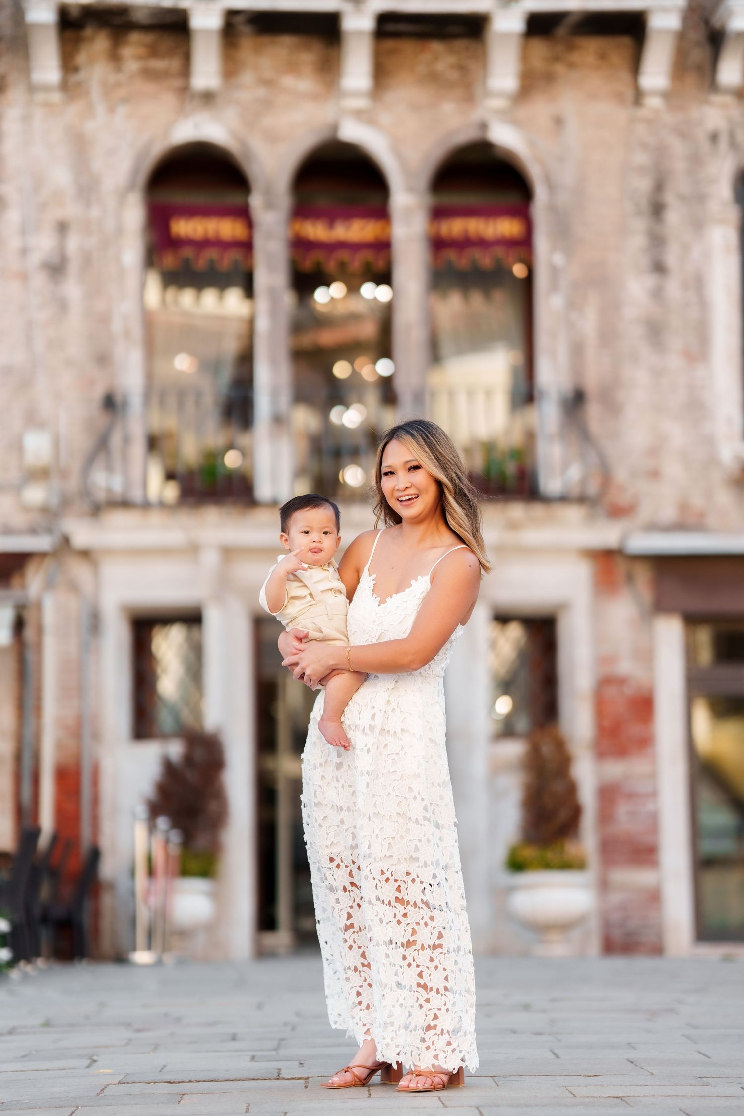 A smiling woman in a white lace dress holds her baby boy in front of a historic Venice building.