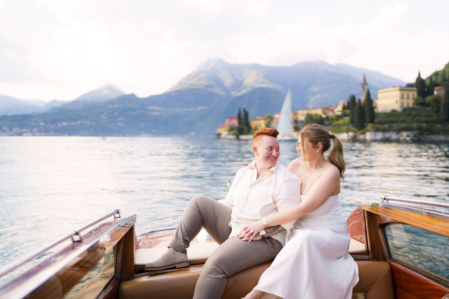 Couple sitting together on a wooden boat on Lake Como, Italy, with mountains and villa in the background.