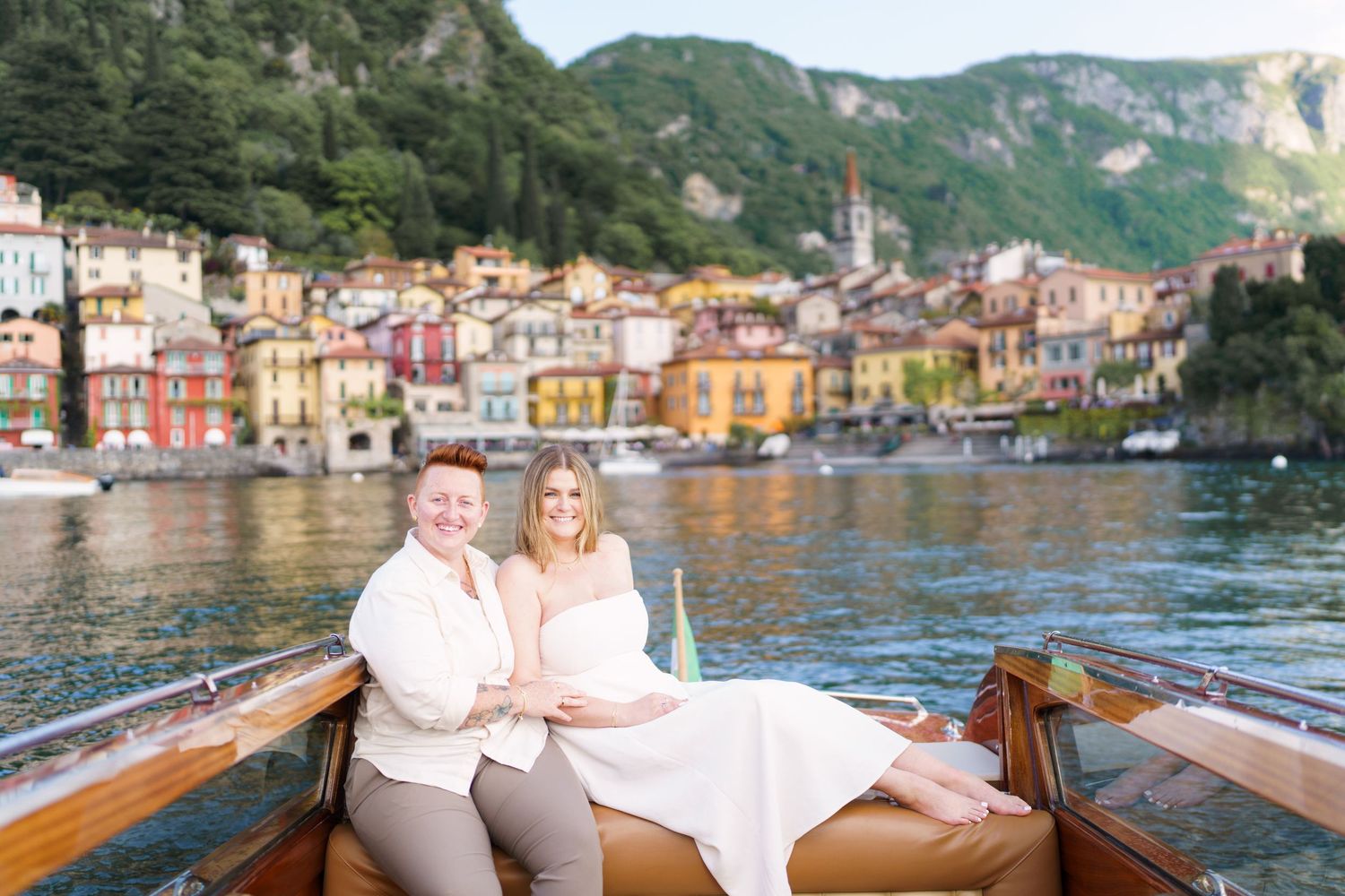 Couple sitting on a wooden boat with colorful Varenna village and Lake Como mountains in the background.
