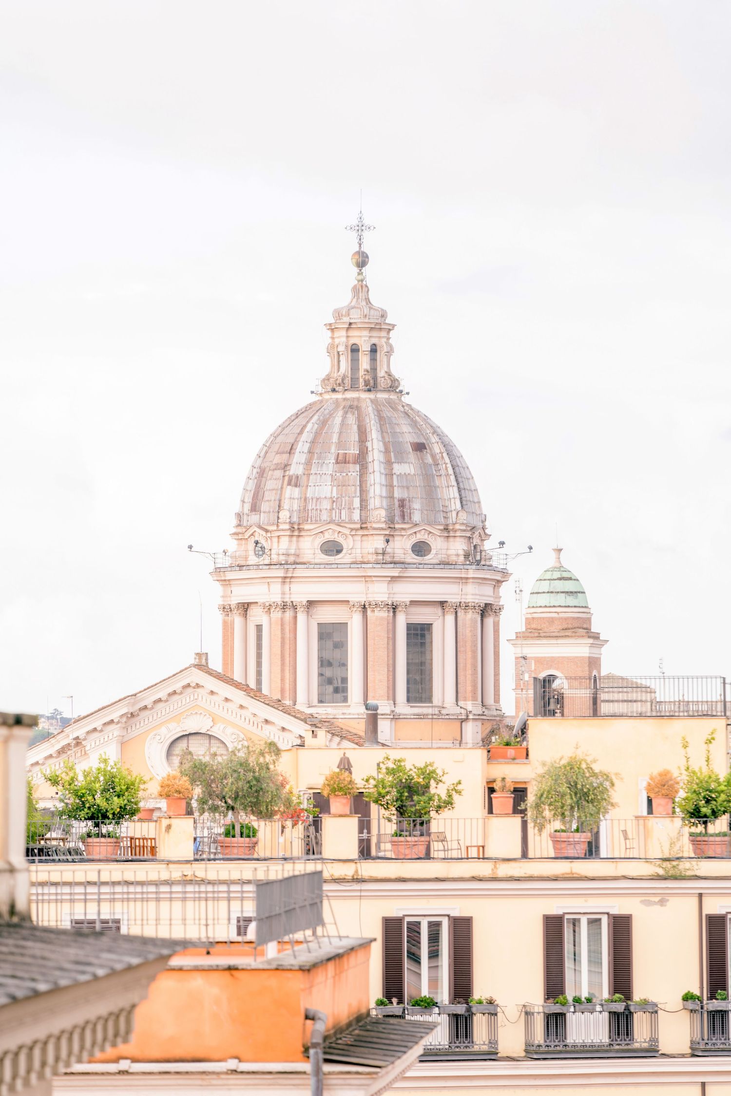 Baroque church dome rises above Roman rooftops with terracotta pots and balconies under a soft overcast sky.