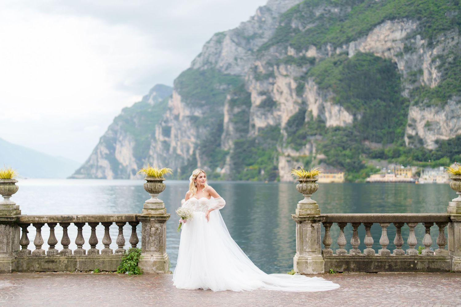 Bride in white gown poses on stone terrace overlooking Lake Garda with dramatic cliffs in background.
