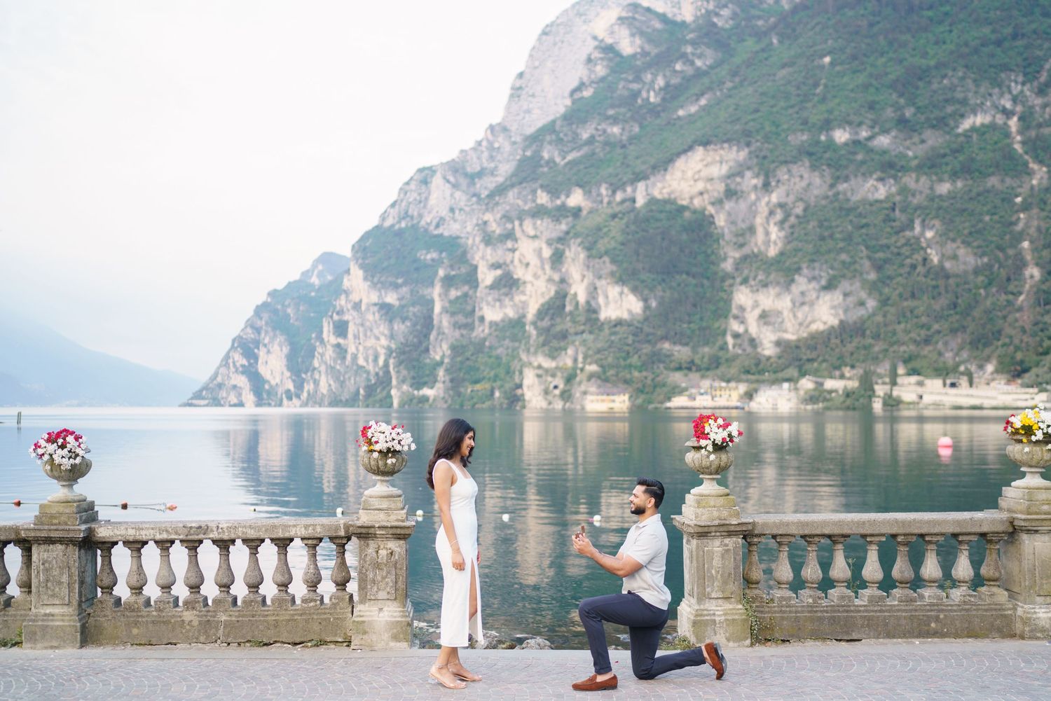 Man proposing to woman on scenic lakeside terrace with mountain backdrop and flower arrangements.