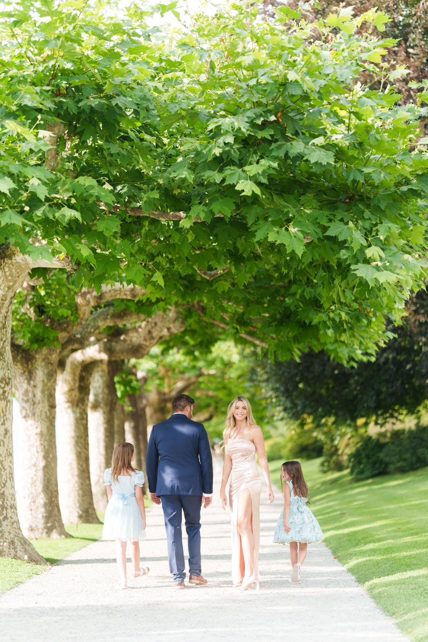 A family walks together along a tree-lined path on a sunny summer day in a lush garden setting.