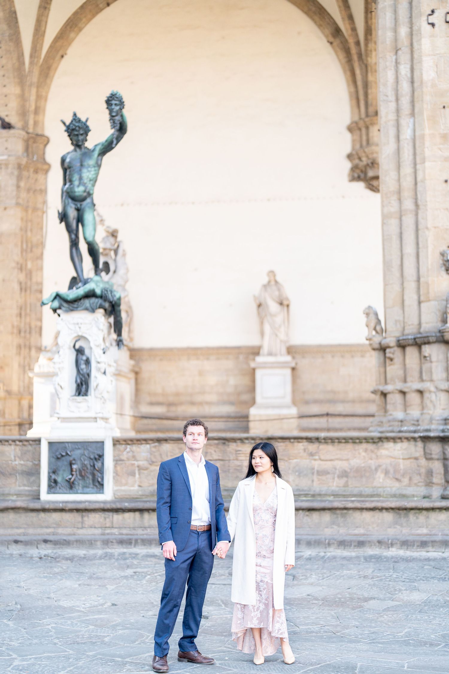Couple poses for engagement photos in front of historic stone archways and bronze statues in Florence, Italy.